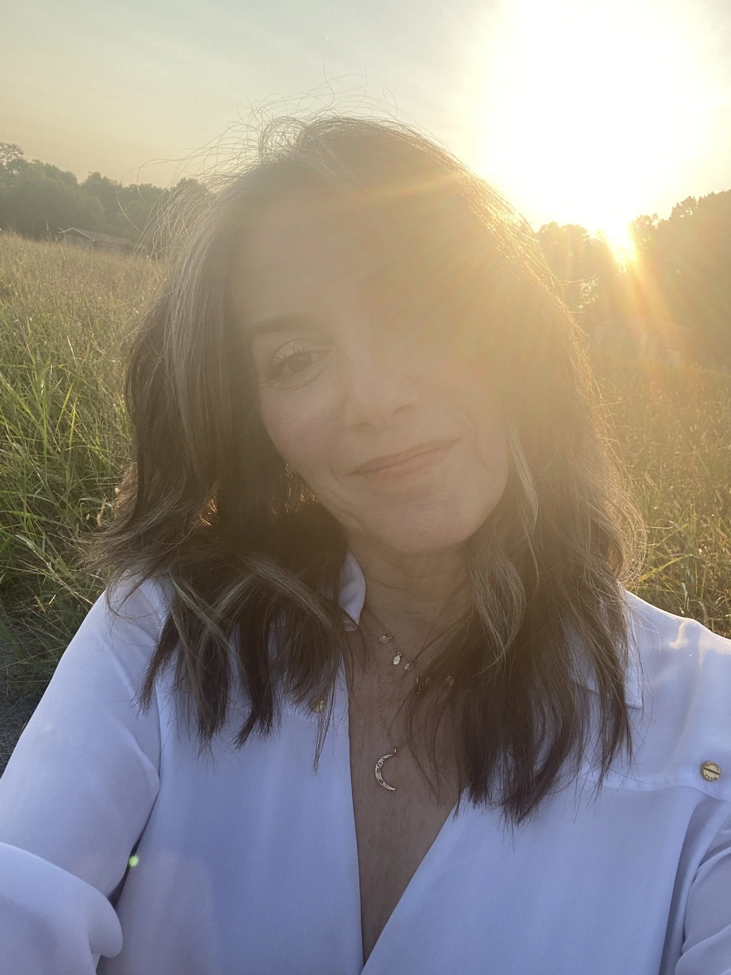 Gina Piccalo, a woman astrologer taking a selfie outdoors in Nashville near the Cumberland River at sunset, with golden sunlight and a field of tall grass in the background.