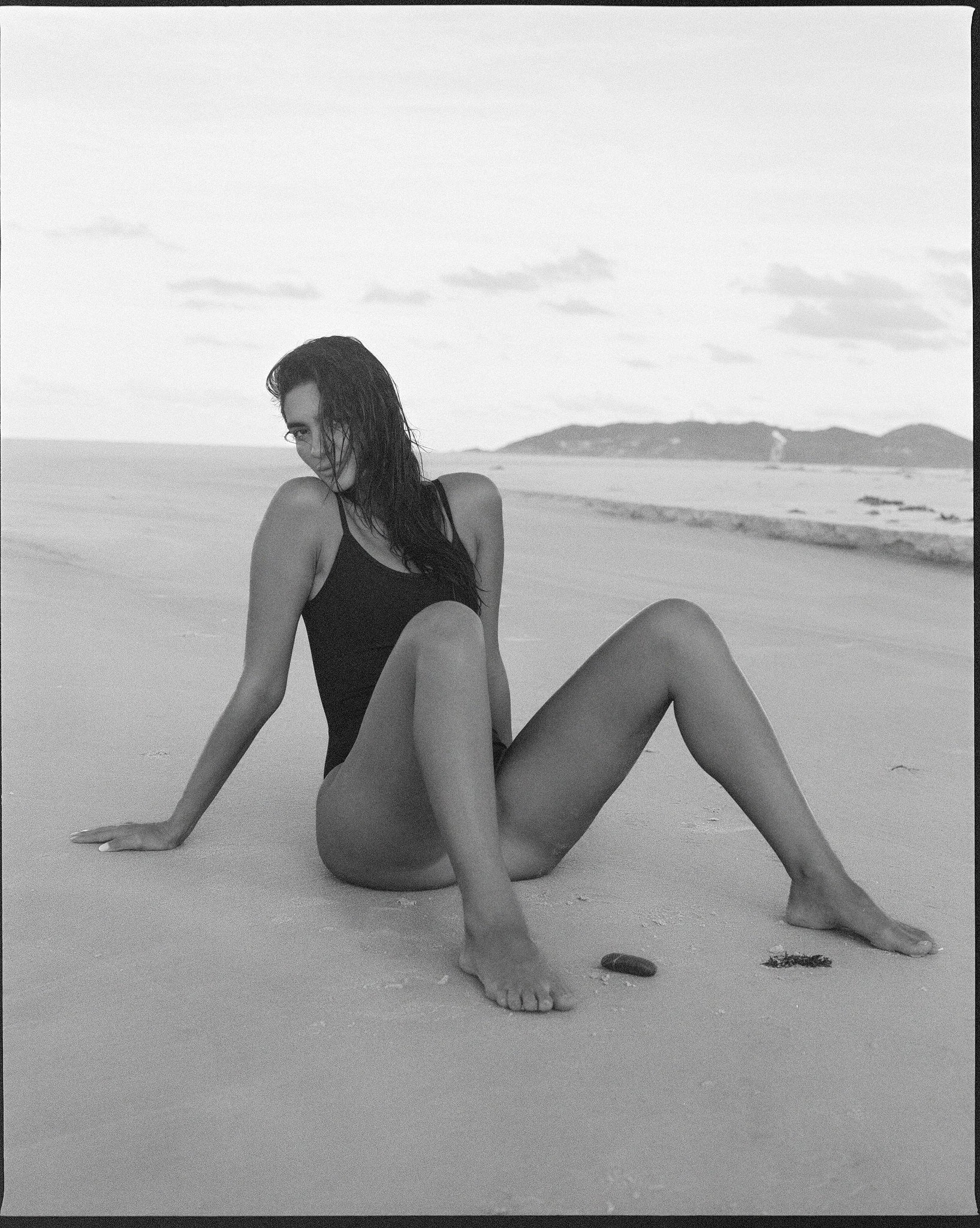 A woman in a black swimsuit sitting on a sandy beach with her legs bent, looking at the camera, with the ocean and distant hills in the background, all in black and white.