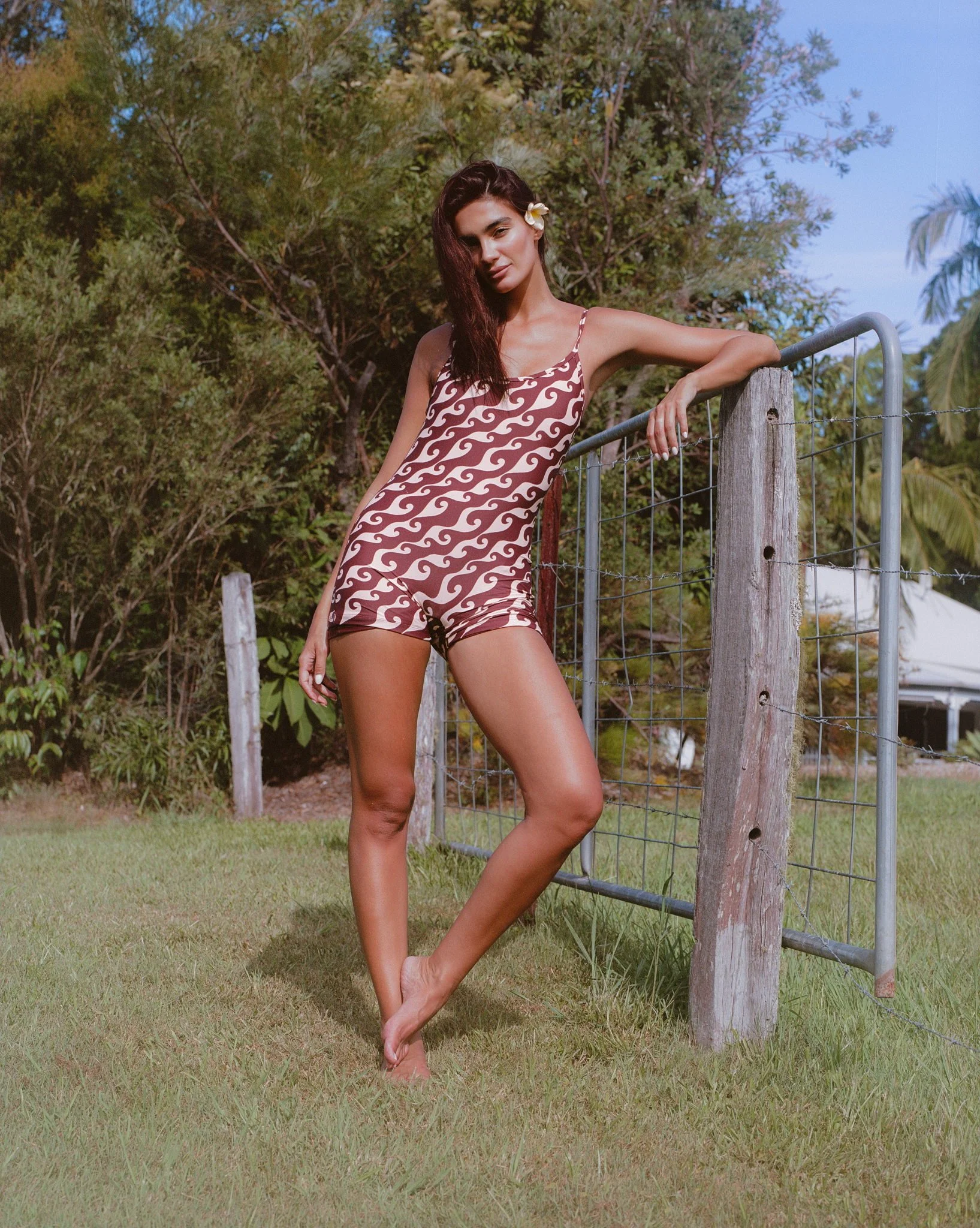 A woman in a brown and white patterned sleeveless romper with spaghetti straps, standing barefoot on grass near a wire fence during daytime, with trees and a blue sky in the background.