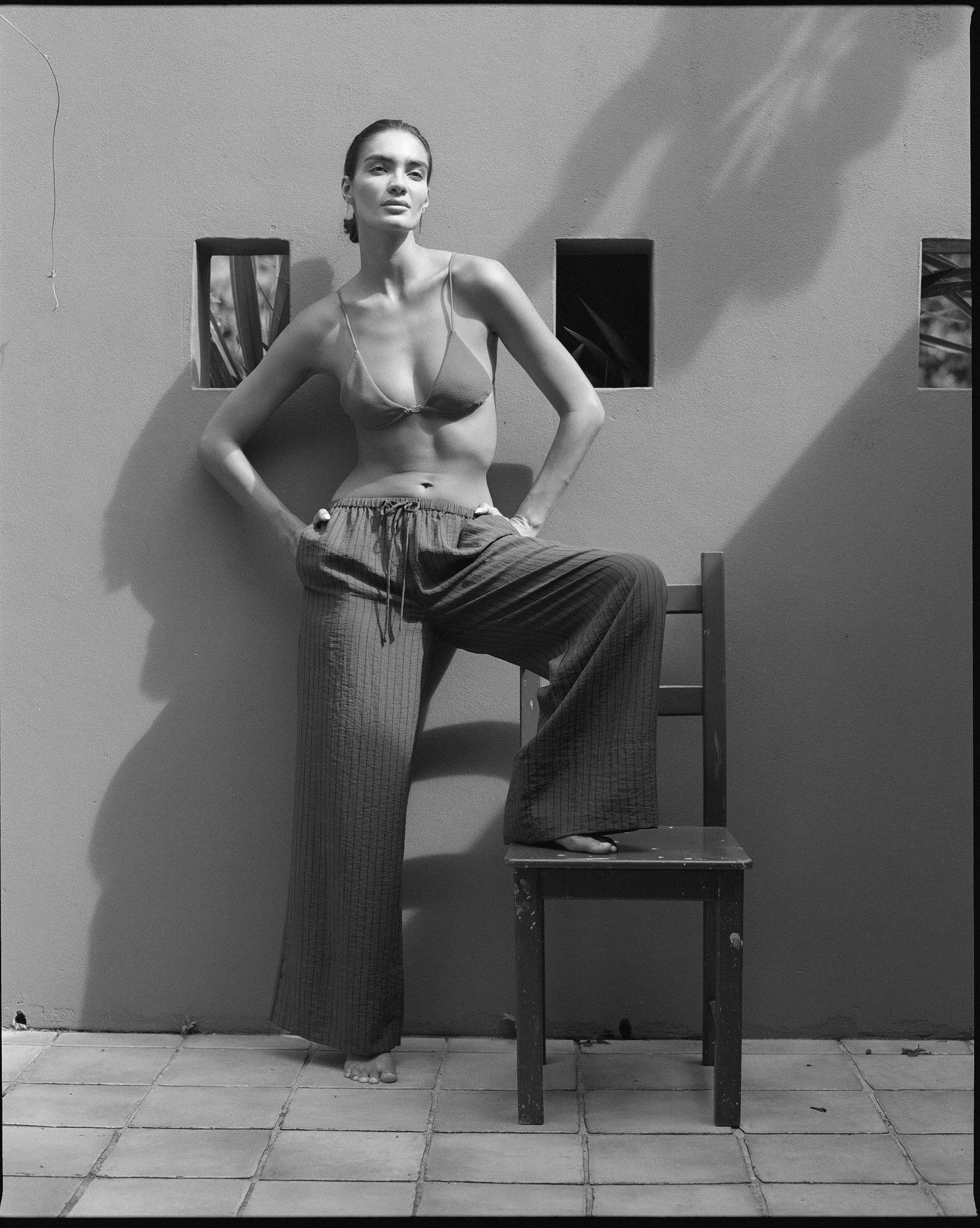 Black and white photo of a woman standing with one foot on a chair. She is wearing a bra and loose pants, standing against a plain wall with small square windows.