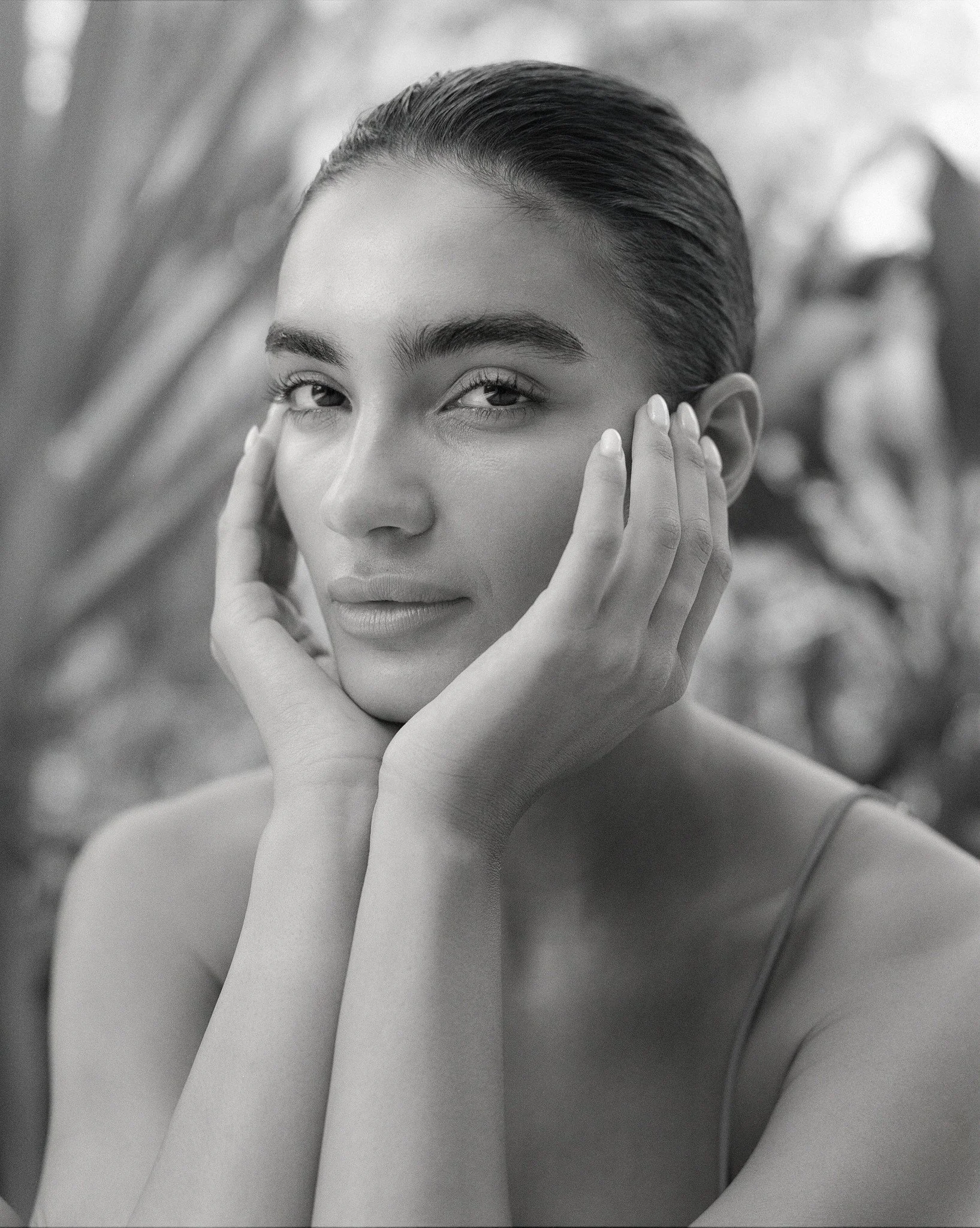 A black-and-white portrait of a woman with her hair slicked back, looking directly at the camera with a confident expression. She is touching her face with both hands, with a blurred natural background.