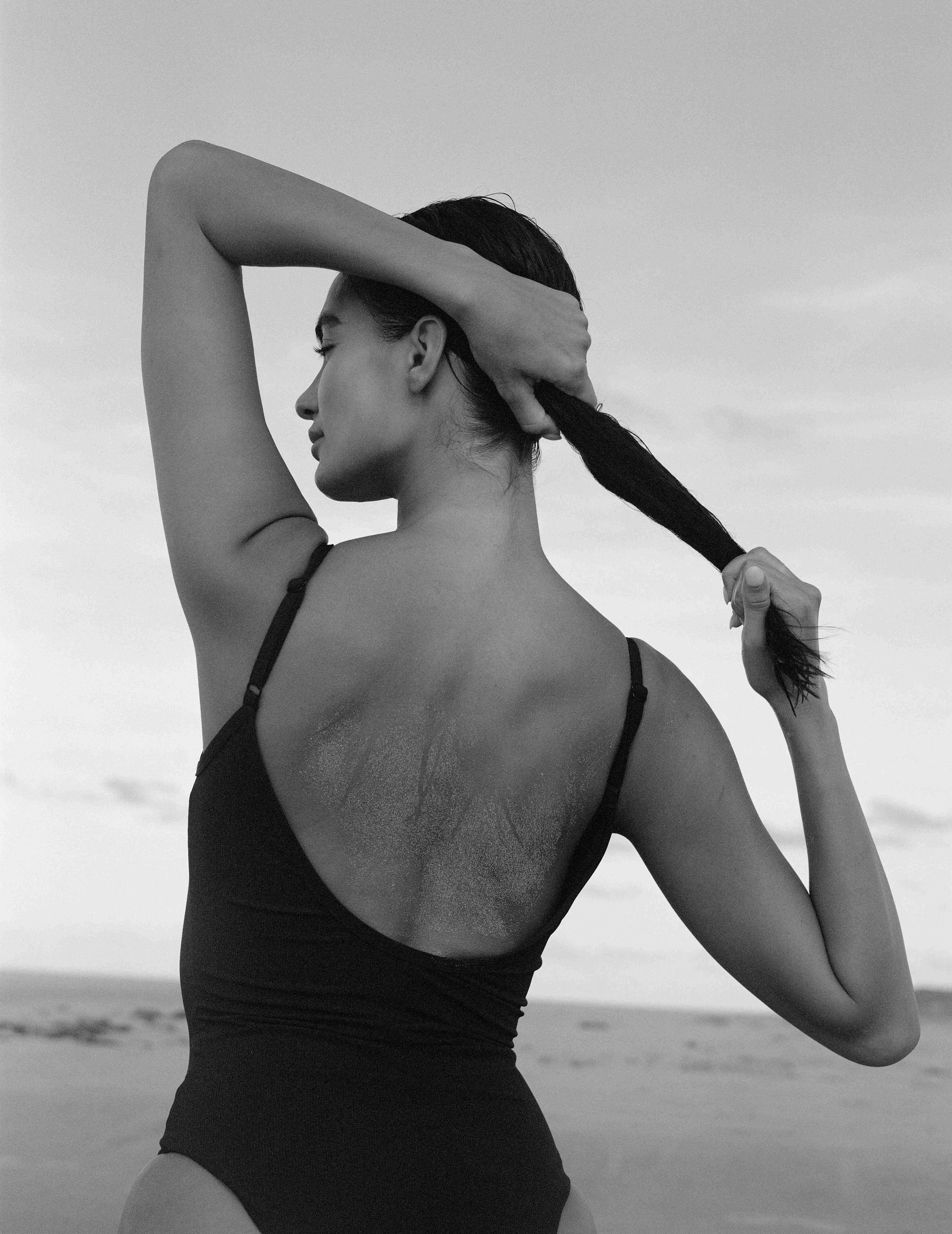 Black and white photo of a woman with long dark hair tying her hair into a ponytail, seen from the back, standing outdoors on a beach.