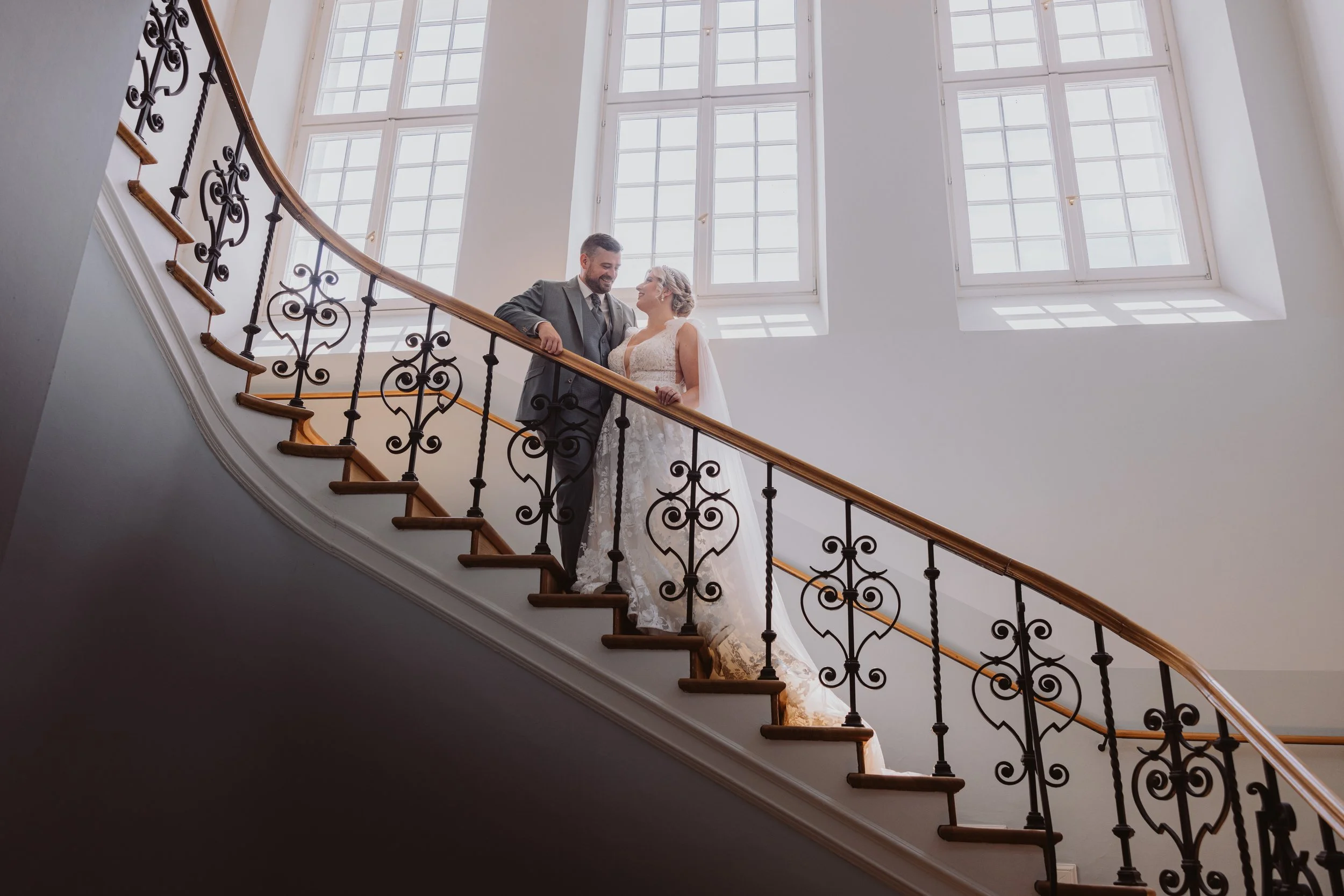 Ein Brautpaar steht auf einer eleganten, geschwungenen Treppe mit kunstvollem schwarzen Geländer, vor großen Fenstern, die viel Tageslicht in den Raum lassen.