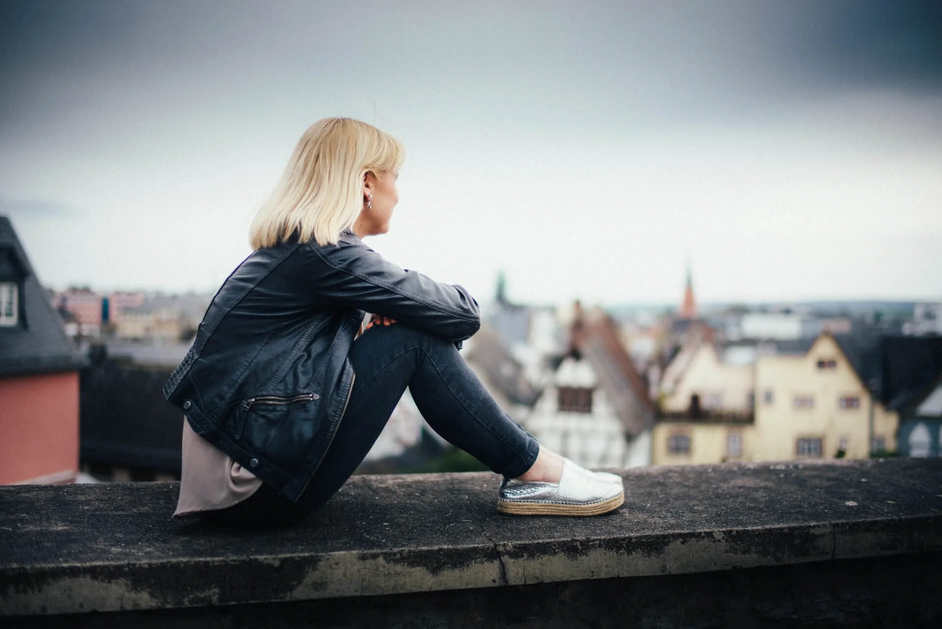 Junge Frau mit blondem Haar in dunkler Lederjacke, sitzend auf einem Dachrand, blickt auf eine Stadt mit alten Häusern.