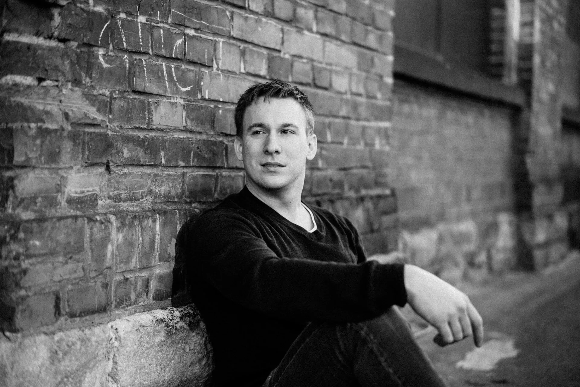 Ein junger Mann sitzt an einer alten Backsteinmauer, schaut nach rechts und wirkt nachdenklich. Das Foto ist schwarz-weiß.