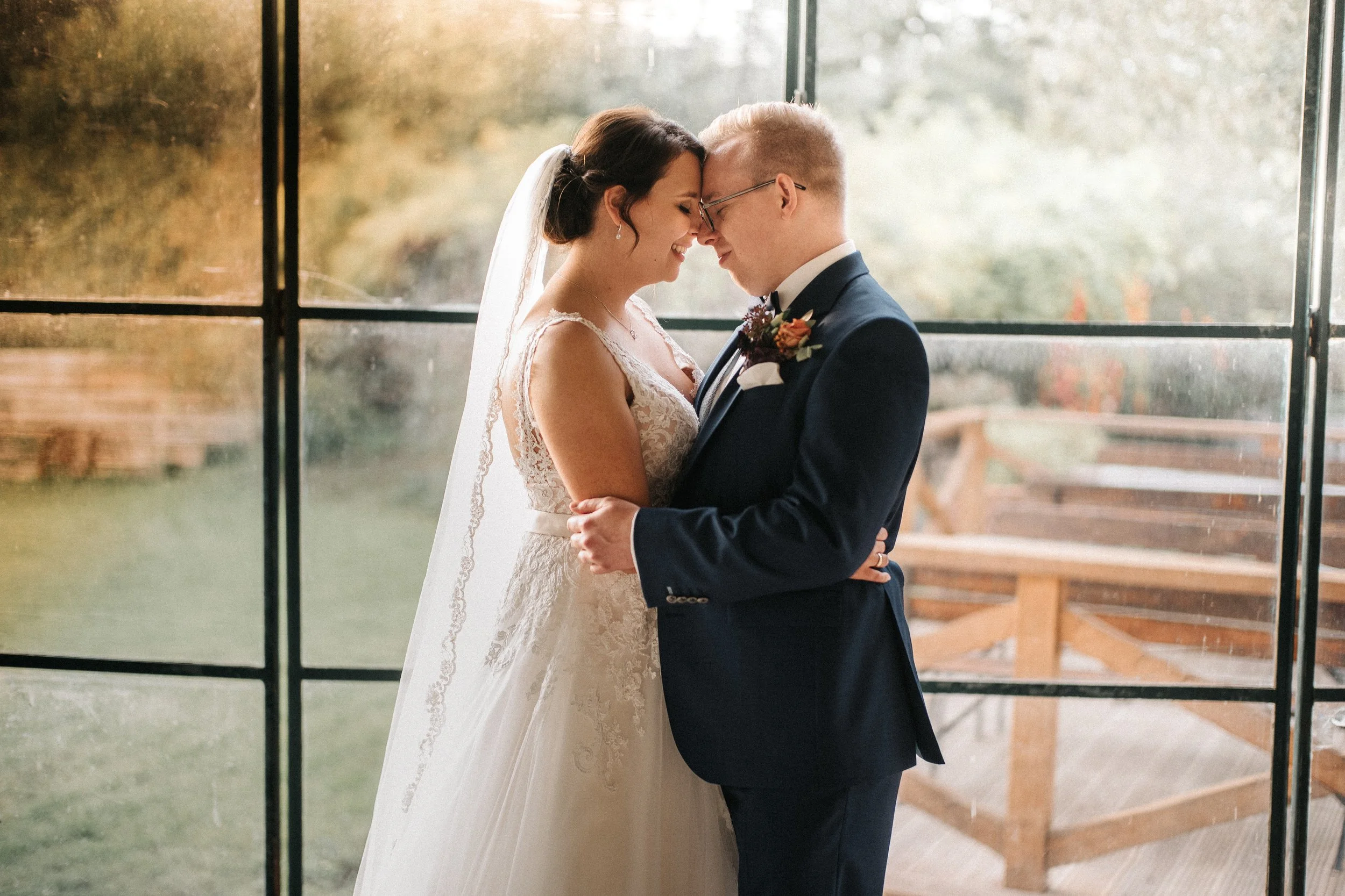 Ein Brautpaar steht sich an einem Fenster gegenüber, lächelt und berührt die Stirn mit den Nasen. Die Braut trägt ein weißes Kleid mit Spitze, die Haare sind elegant hochgesteckt, und sie trägt einen Schleier. Der Bräutigam trägt einen schwarzen Anzu