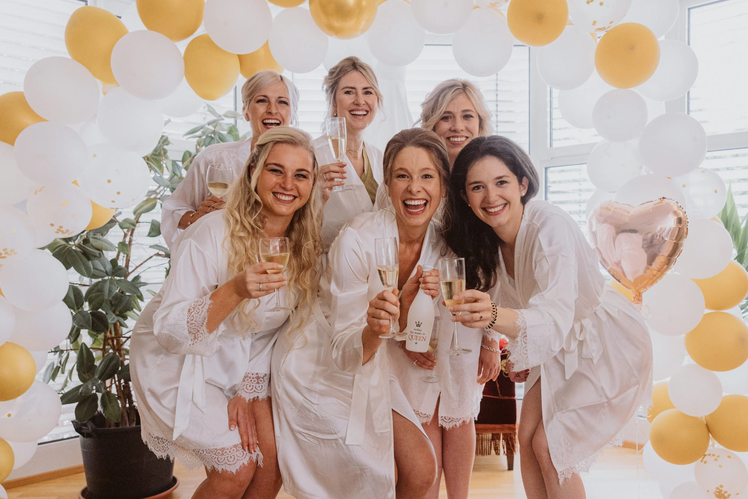 Eine Gruppe von Frauen in weißen Nachthemden, die bei einer Feier mit Sekt auf einem Hintergrund aus weißen und goldenen Ballons lachen und anstoßen.