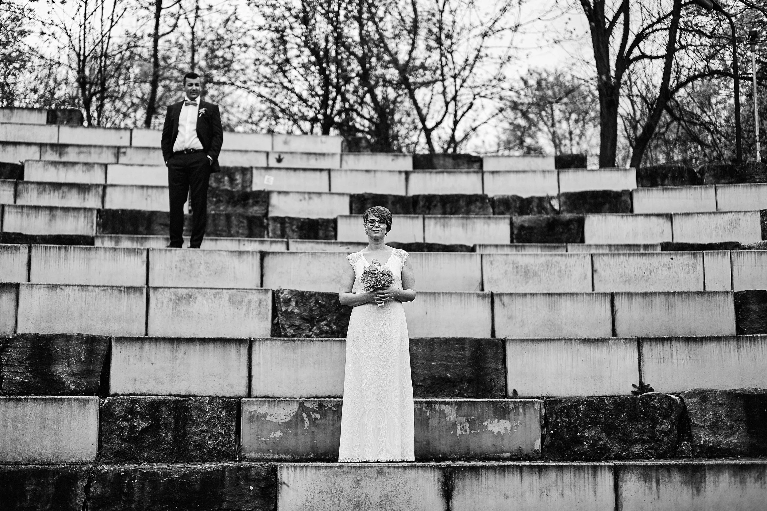 Ein Brautpaar steht auf einer alten, abgenutzten Treppe im Freien. Die Frau trägt ein langes, elegantes Hochzeitskleid und hält einen Blumenstrauß. Der Mann trägt einen dunklen Anzug mit Fliege. Es ist in Schwarzweiß gehalten, mit Bäumen im Hintergru