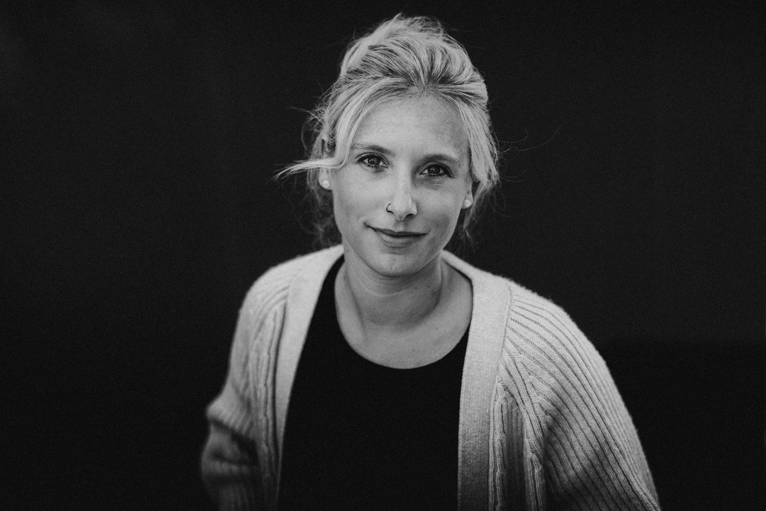 Schwarz-weiß-Porträt einer Frau mit lockigem, blonden Haar, einem Nasenring und einem freundlichen Lächeln, getragen von einem grauen Pullover und einem schwarzen Shirt, vor dunklem Hintergrund.
