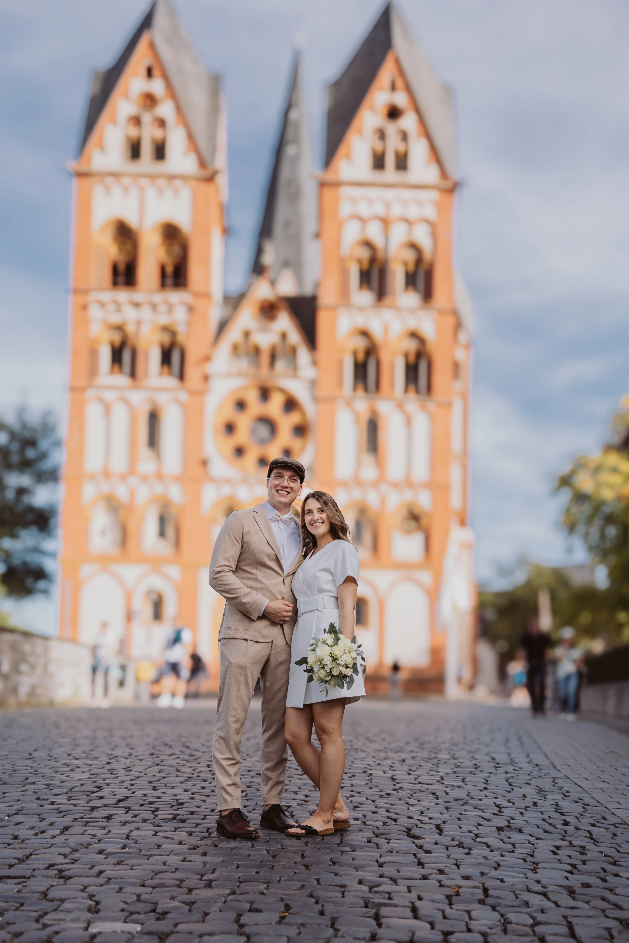 Ein Paar steht lachend vor einer Kirche mit Türmen, das Paar trägt formelle Kleidung, der Mann in einem Anzug, die Frau in einem weißen Kleid mit Blumenstrauß.