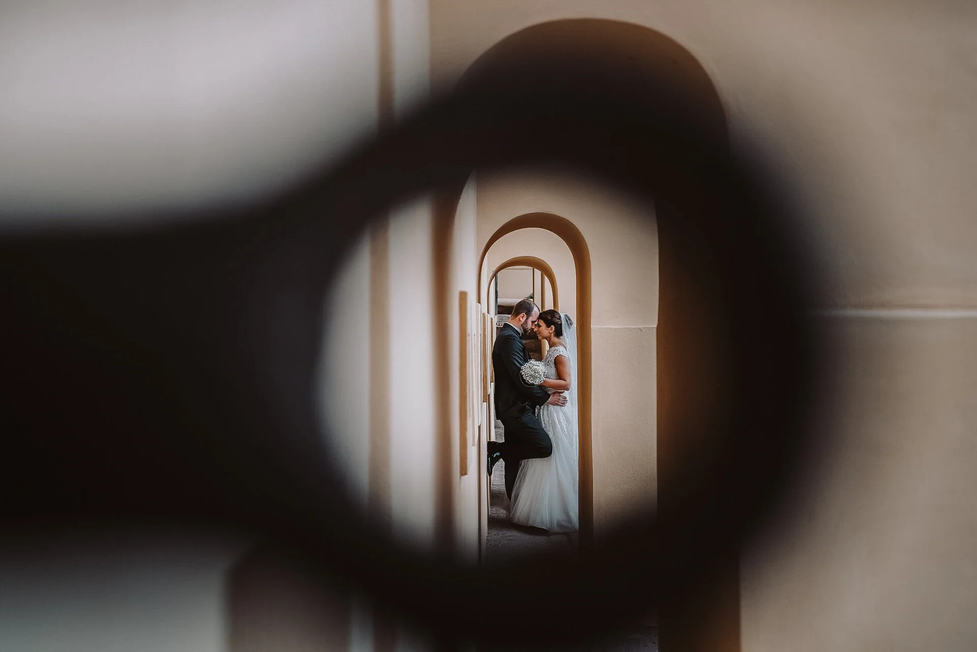 Braut und Bräutigam, die sich an einer Wand in einem Flur, durch eine kleine Öffnung fotografiert, küssen.
