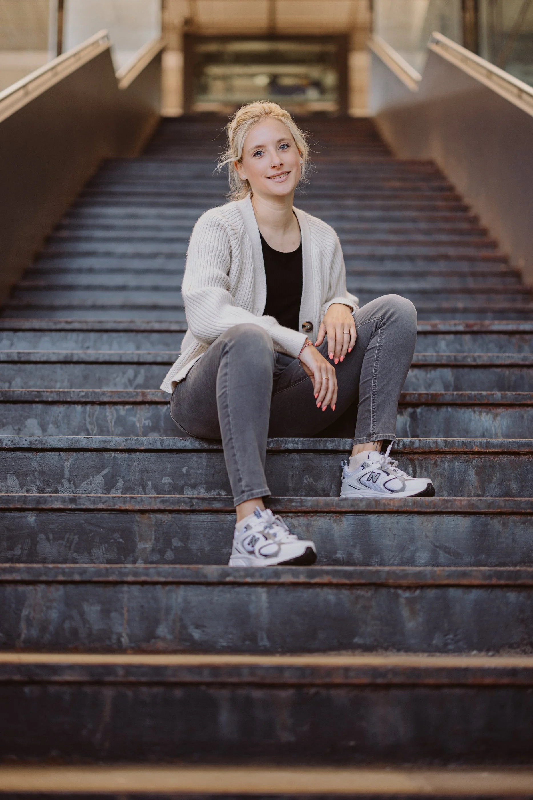 Eine junge Frau sitzt auf einer Treppe aus Metall, trägt graue Jeans, weiße Turnschuhe, ein schwarzes Oberteil und eine weiße Strickjacke, lächelt und schaut in die Kamera.