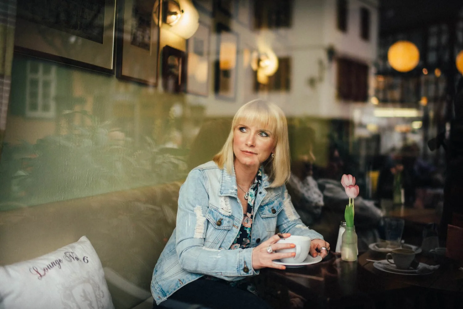 Eine blonde Frau in einem Jeansjacke sitzt in einem Café und hält eine Tasse Kaffee. Im Hintergrund sind andere Gäste und die Einrichtung des Cafés sichtbar.