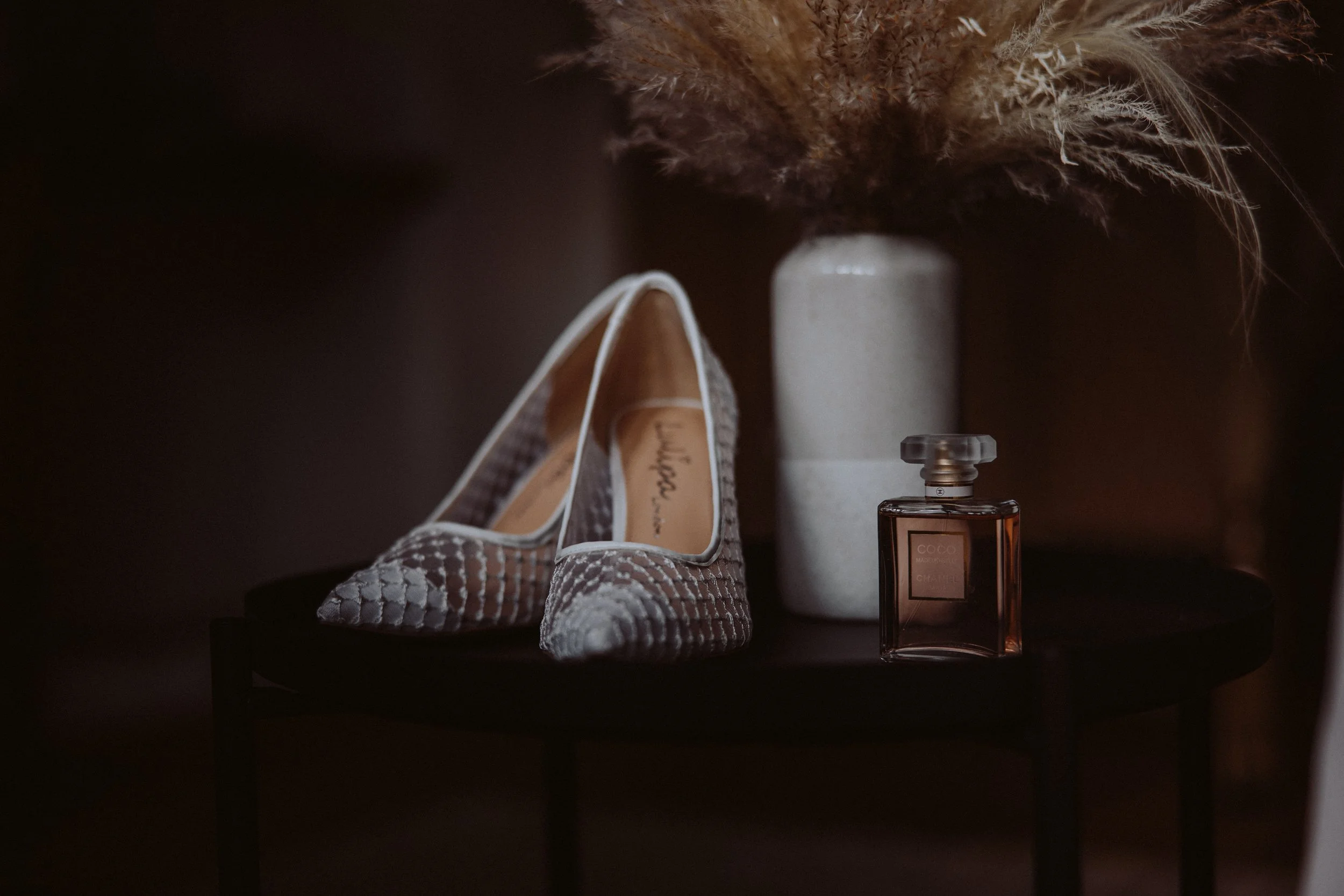 Paar High Heels, einen Parfümflakon, eine weiße Vase und eine Vase mit Trockenblumen auf einem schwarzen runden Beistelltisch.