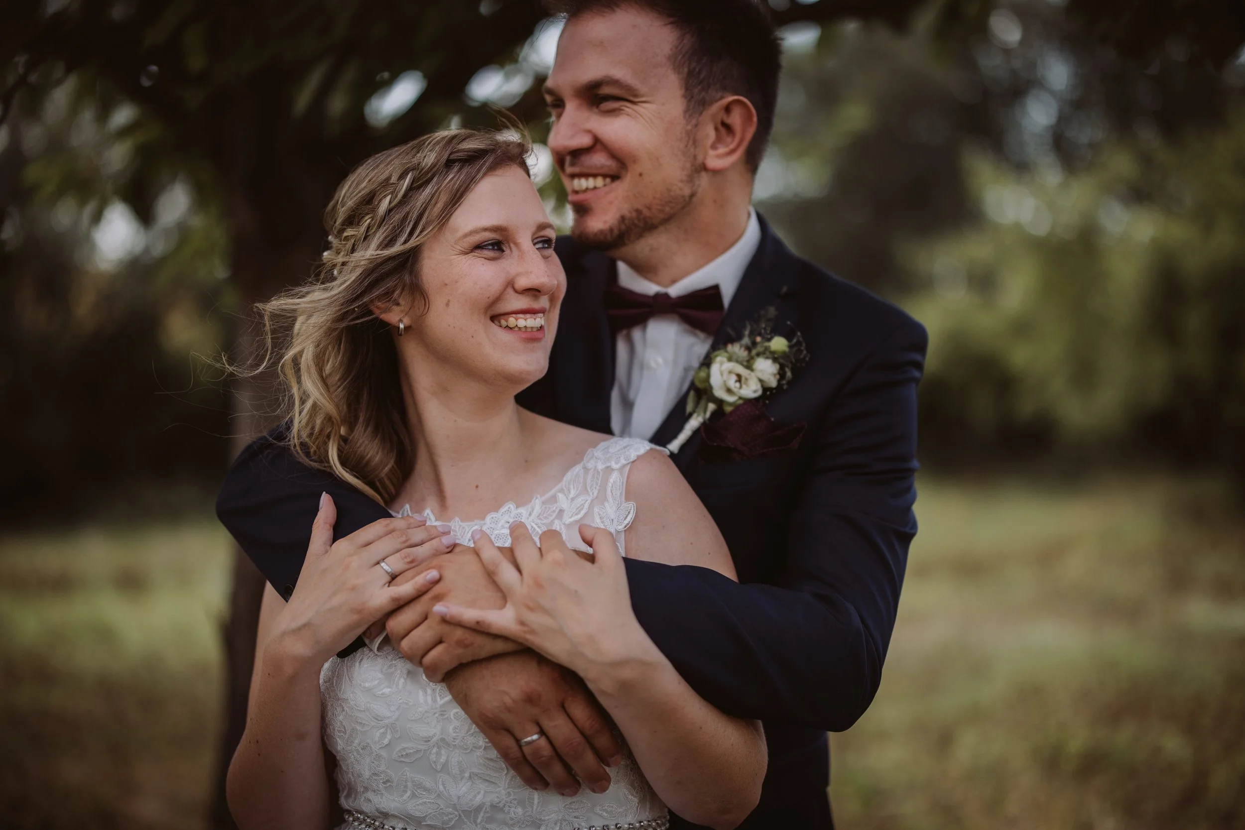 Ein glückliches Brautpaar beim Fotoshooting im Freien, umgeben von Bäumen, lächelnd und umarmend.