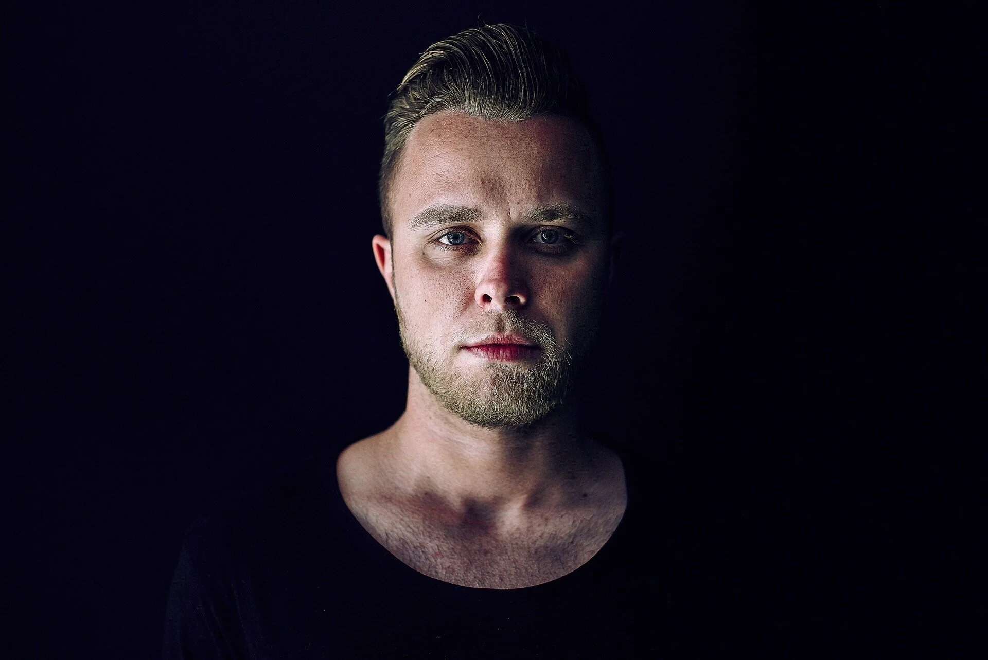 Porträt eines jungen Mannes mit blondem Haar und Bart vor schwarzem Hintergrund.