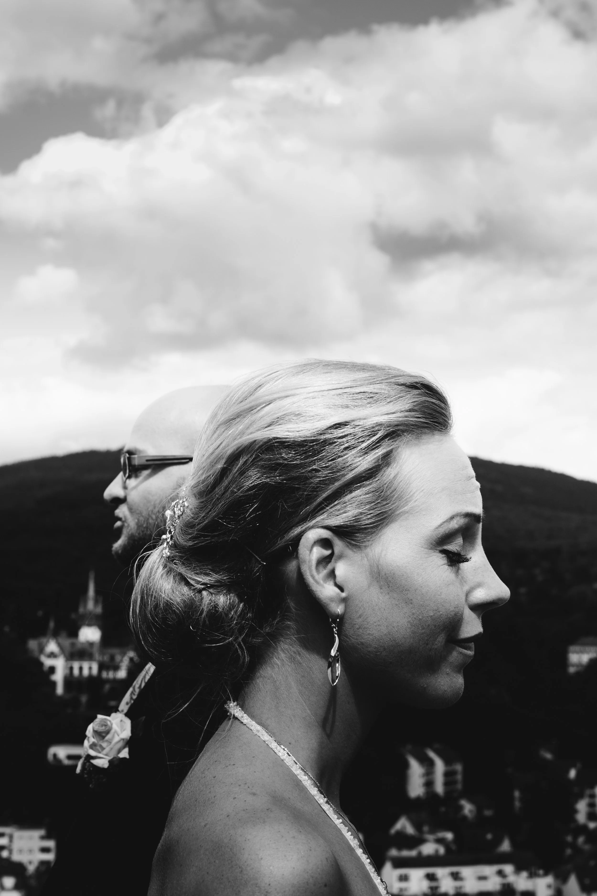 Schwarz-Weiß-Foto eines Paares, bei dem die Frau im Vordergrund mit seitlichem Profil und der Mann im Hintergrund mit frontalem Profil zu sehen ist. Beide haben geschlossene Augen und tragen elegante Kleidung, im Hintergrund sind Häuser und Hügel bei
