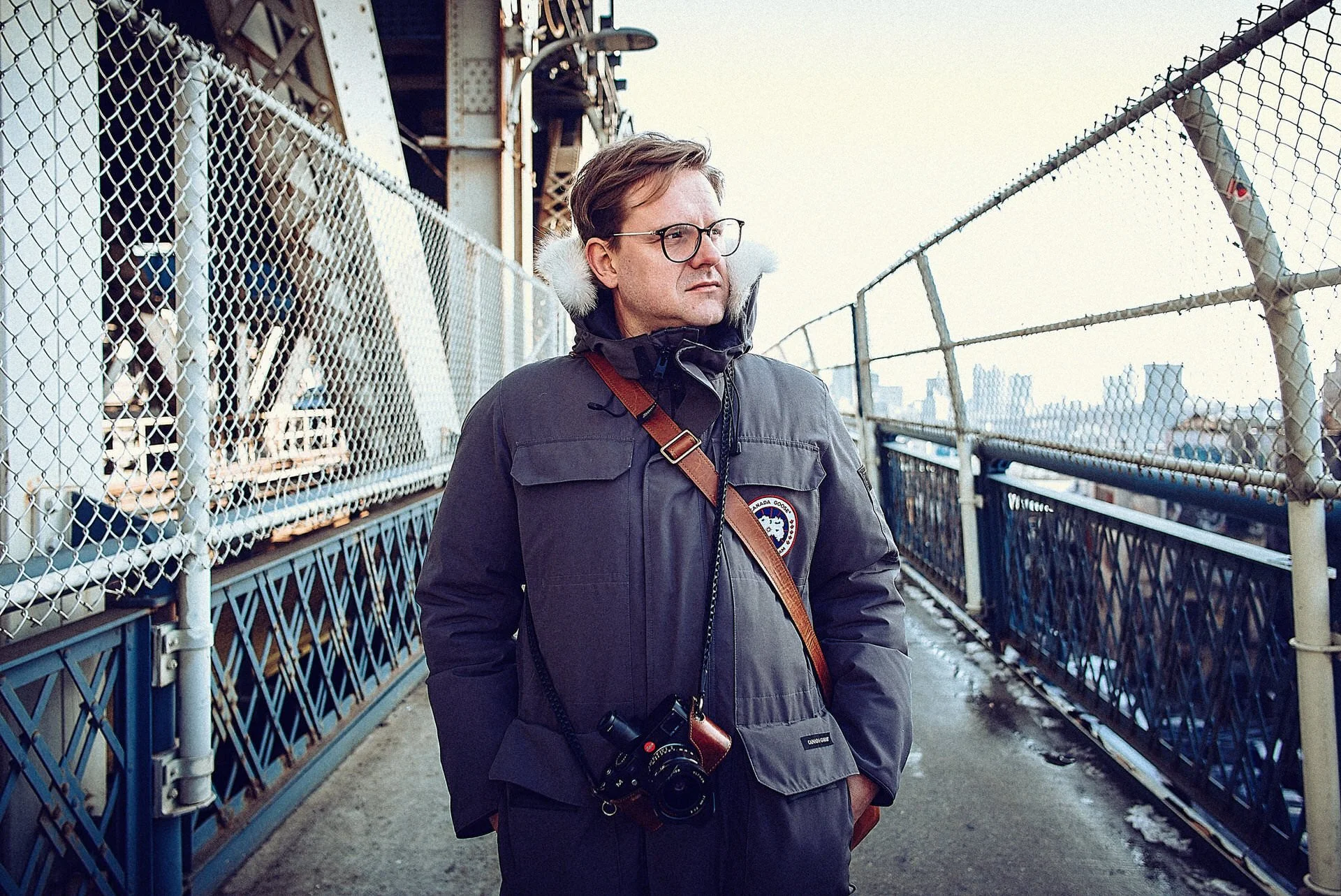 Ein Mann steht auf einer Brücke, trägt eine dunkle Jacke mit einer Patch auf der Brust, eine Kamera um den Hals, trägt Brille und hat kurzbraune Haare, während er in die Ferne blickt.