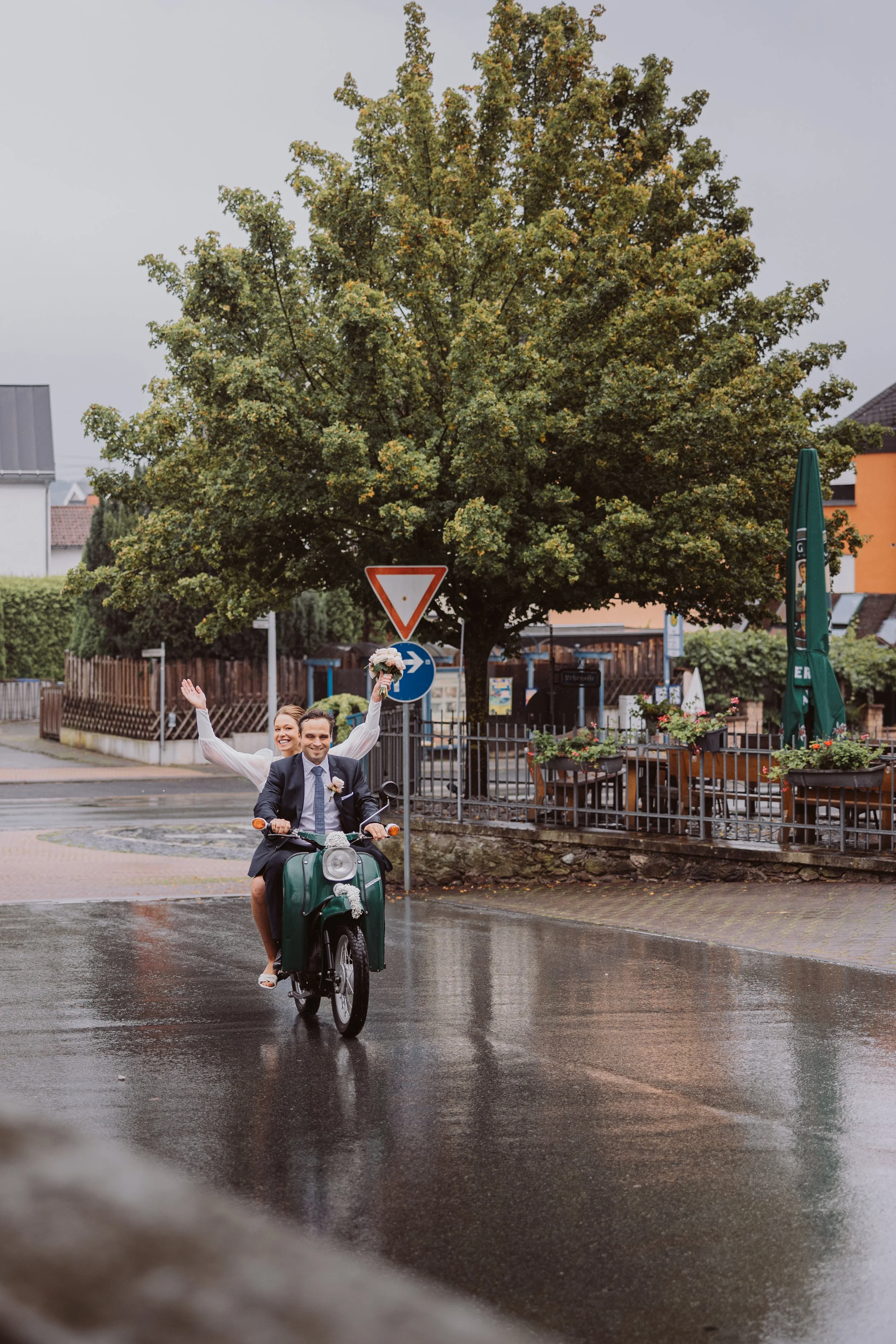 Ein lachendes Paar in formeller Kleidung fährt auf einem Motorrollerscooter im Regen, die Frau hält einen Blumenstrauß hoch, bei Regen auf einer Straße mit großem Baum im Hintergrund.