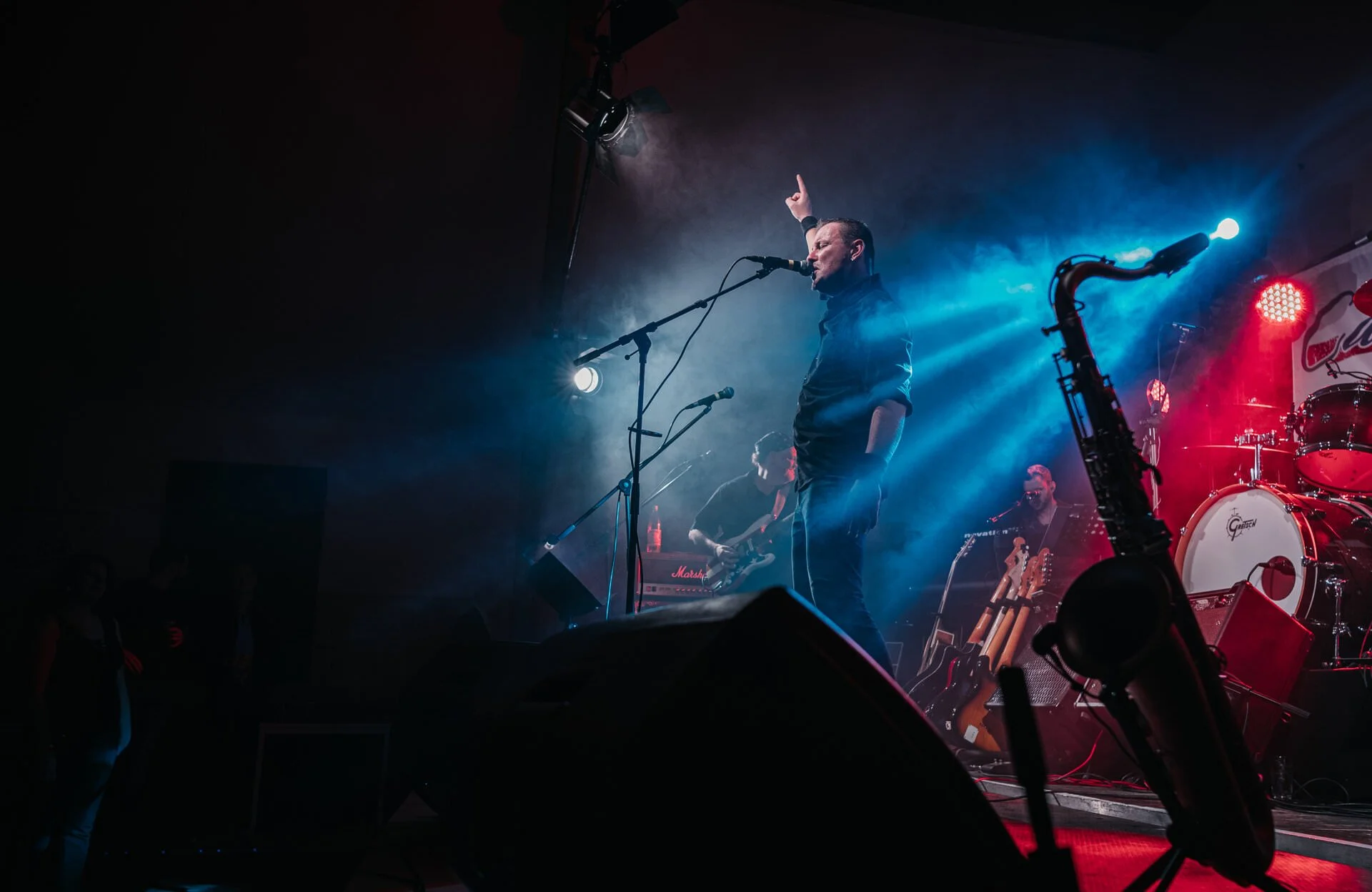 Ein Musiker steht auf der Bühne und singt in ein Mikrofon, umgeben von anderen Bandmitgliedern und Musikinstrumenten, mit buntem Bühnenlicht im Hintergrund.