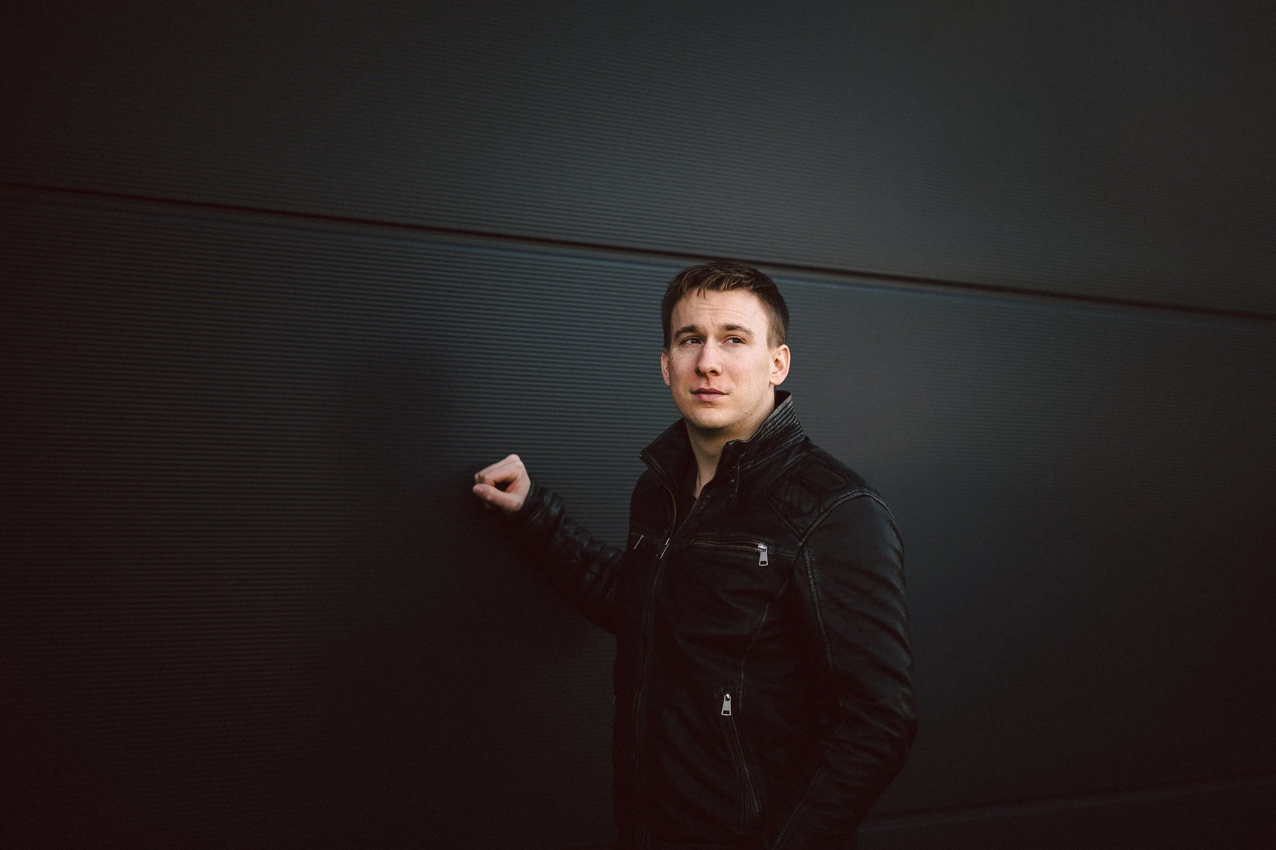 Junger Mann in schwarzer Lederjacke lehnt an einer dunklen Wand.