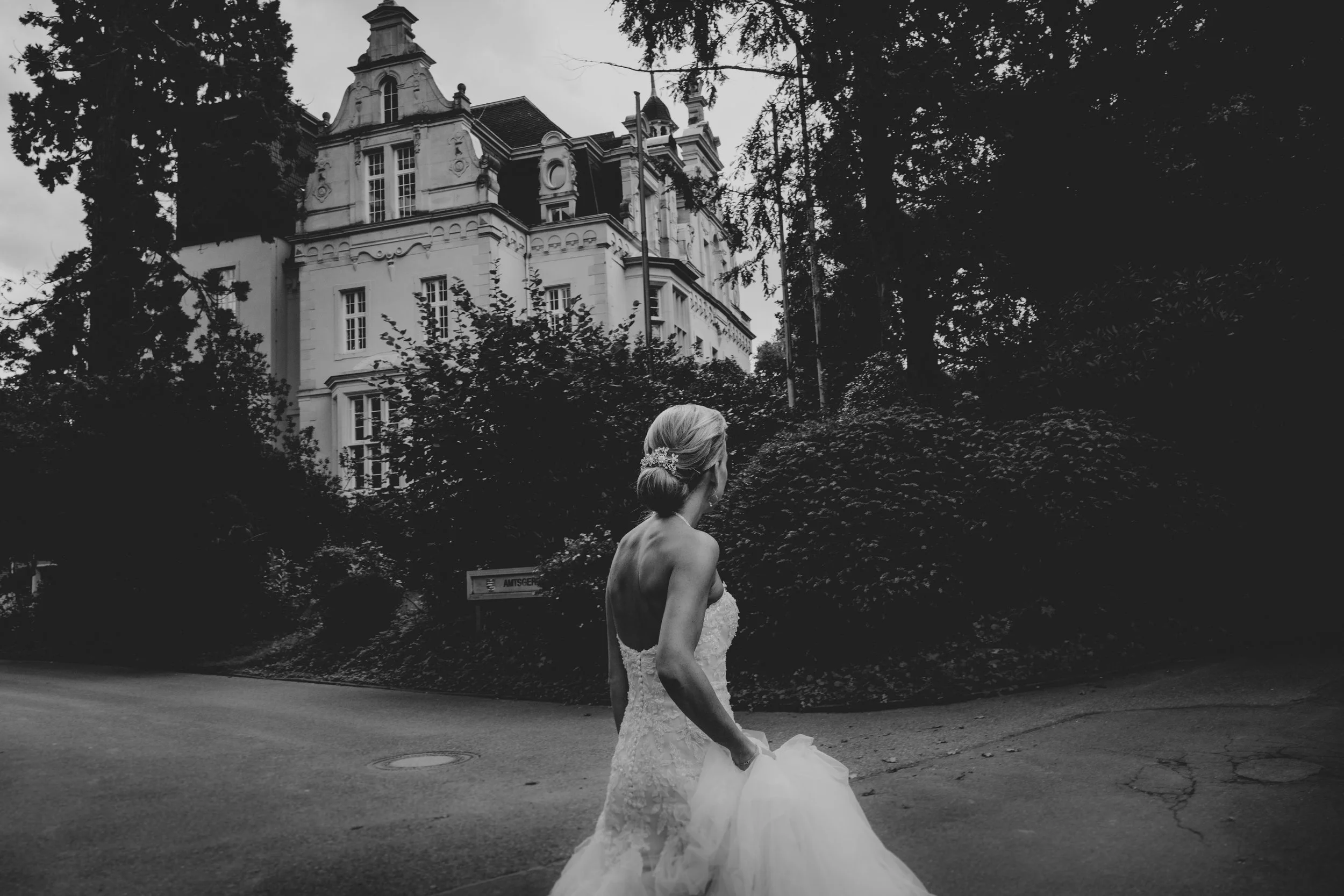 Eine Braut in einem weißen Hochzeitskleid läuft auf einer Straße in einer parkähnlichen Umgebung, im Hintergrund ein großes historisches Gebäude und Bäume.