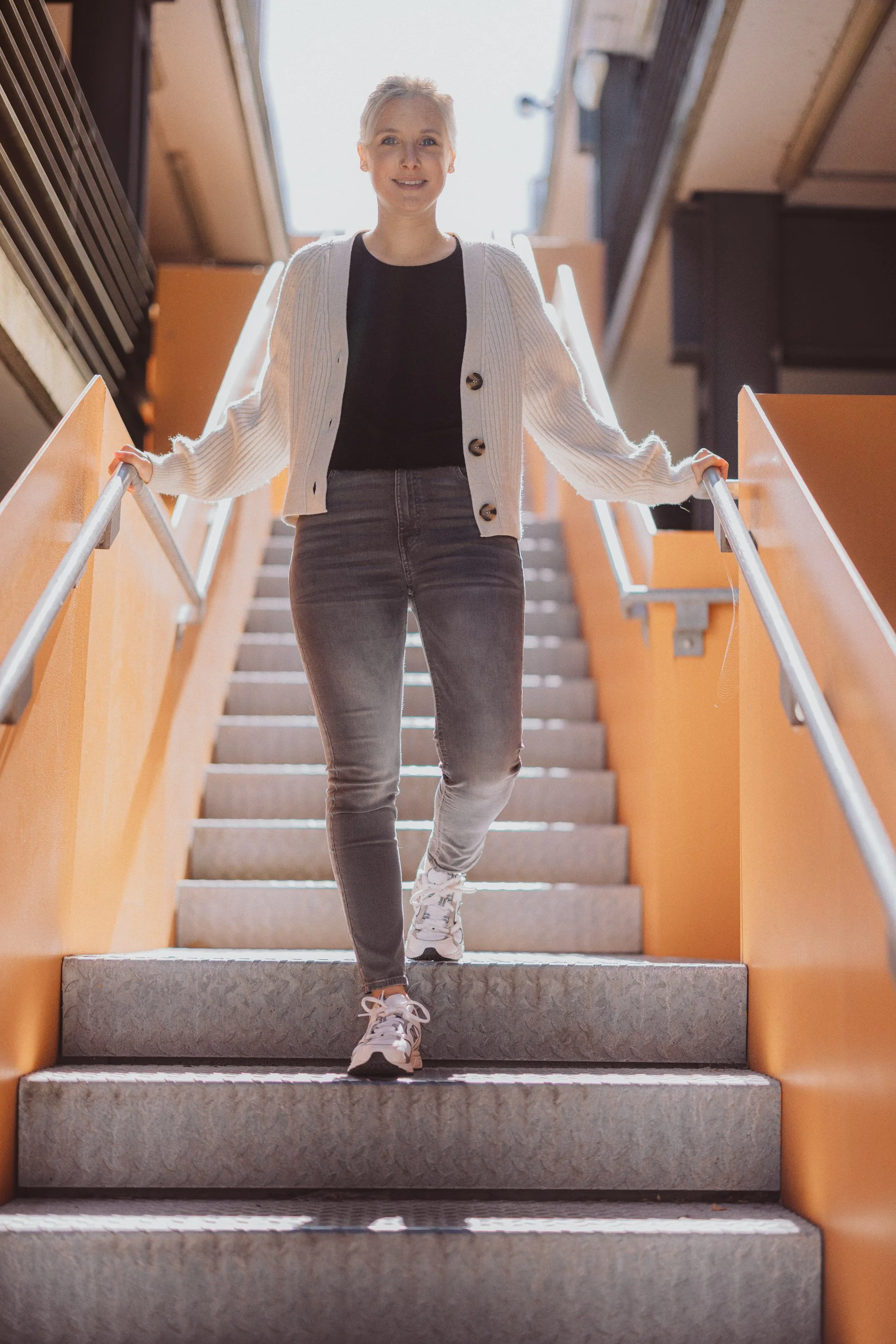 Eine junge Frau mit grau-blonden Haaren, die eine Treppe hinuntergeht, trägt ein schwarzes T-Shirt, eine weiße Strickjacke, graue Jeans und Turnschuhe, während sie nach unten schaut und lächelt.