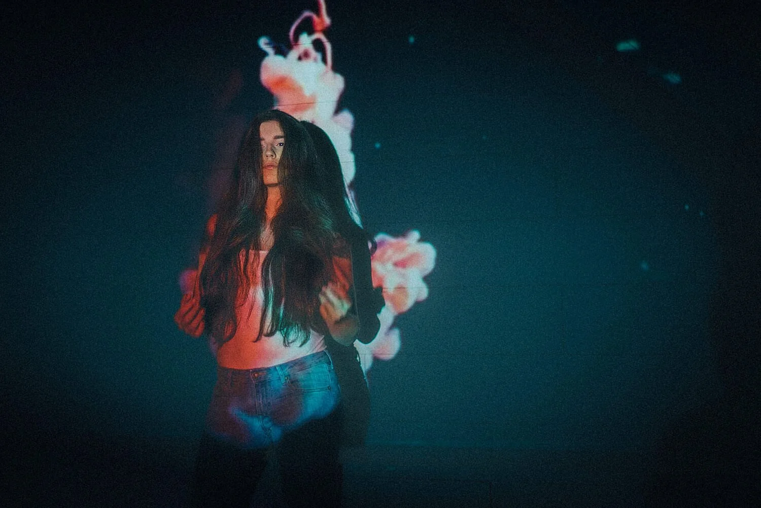 Junge Frau mit langen dunklen Haaren steht vor einer dunklen Hintergrundprojektion mit bunten, nebligen Wolkenmustern, trägt ein weißes Oberteil und Jeans.