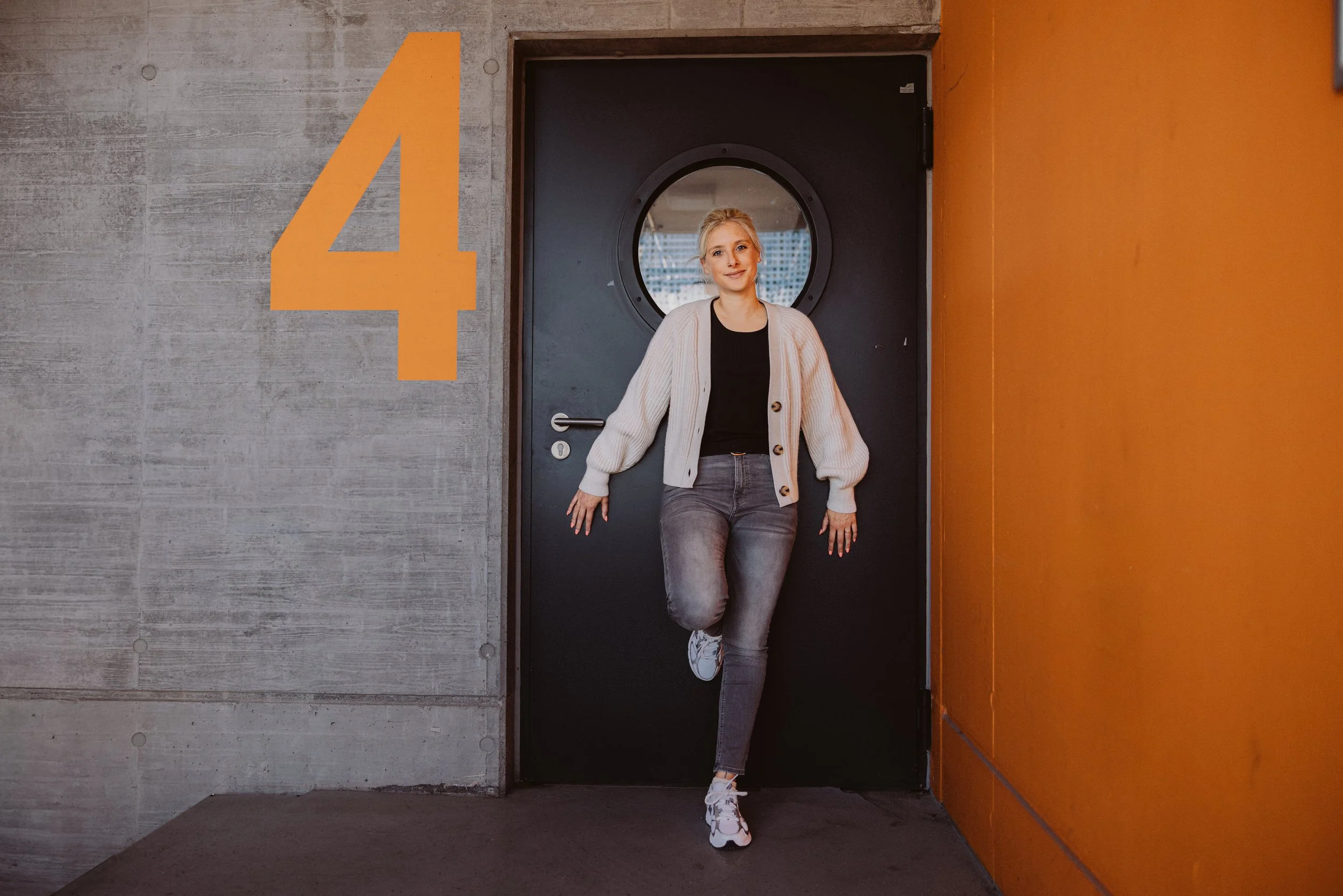 Junge Frau in einem Flur lehnt an einer schwarzen Tür, daneben eine Wand mit der großen orangefarbenen Zahl 4, an der rechten Seite eine orange Wand.