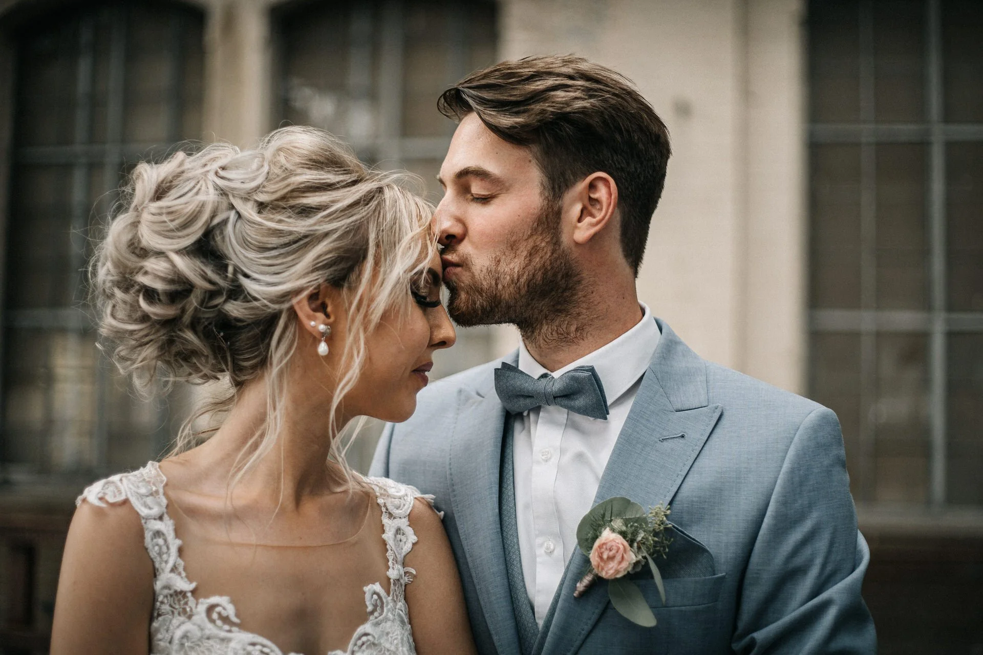 Ein Brautpaar küsst sich bei einer Hochzeit, der Mann trägt einen hellblauen Anzug mit Fliege und die Frau trägt ein weißes Hochzeitskleid mit Spitze.