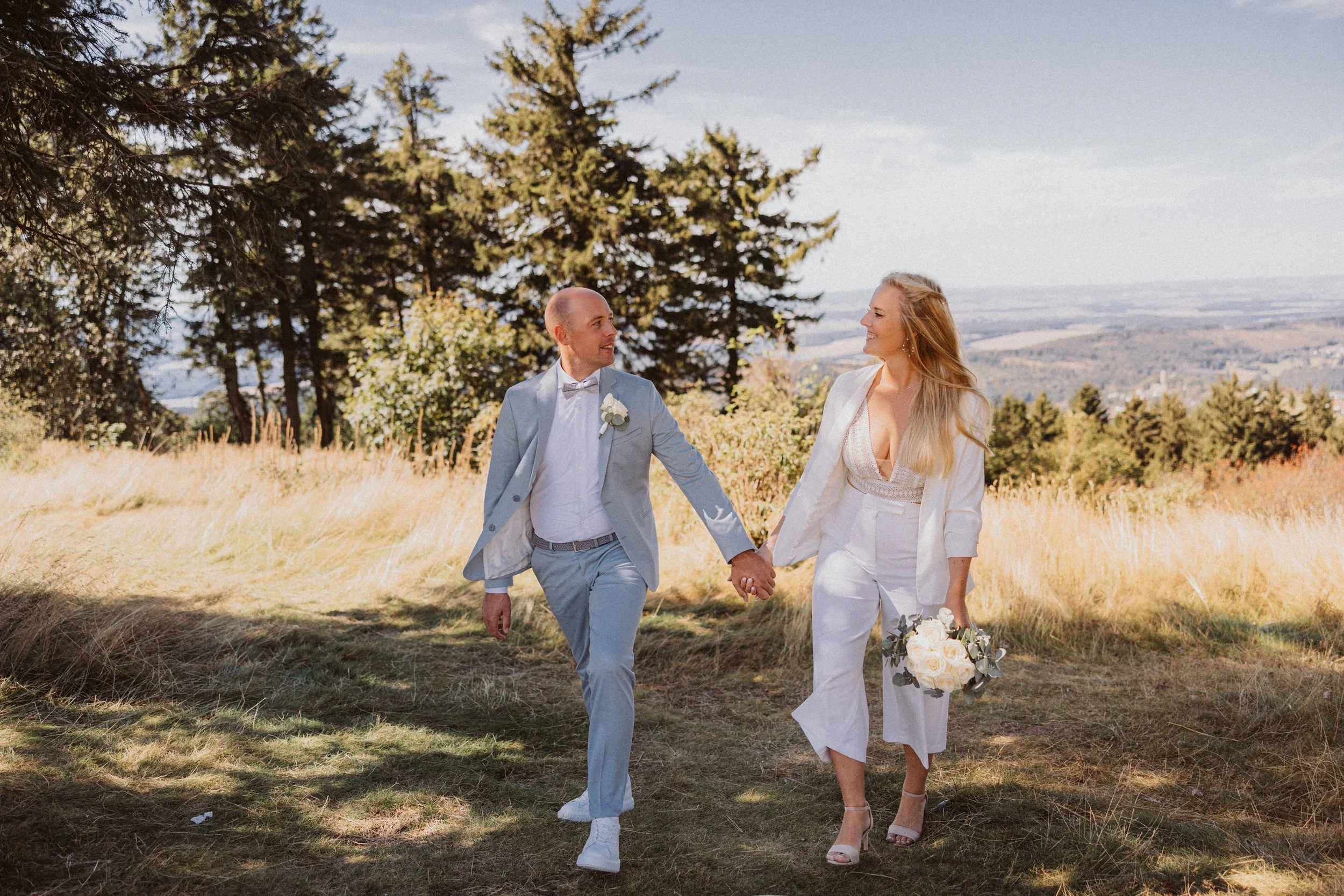 Ein Mann und eine Frau in eleganter Kleidung beim Spaziergang im Freien, die Hand in Hand halten, im Hintergrund Bäume und eine Landschaft, sonniges Wetter.
