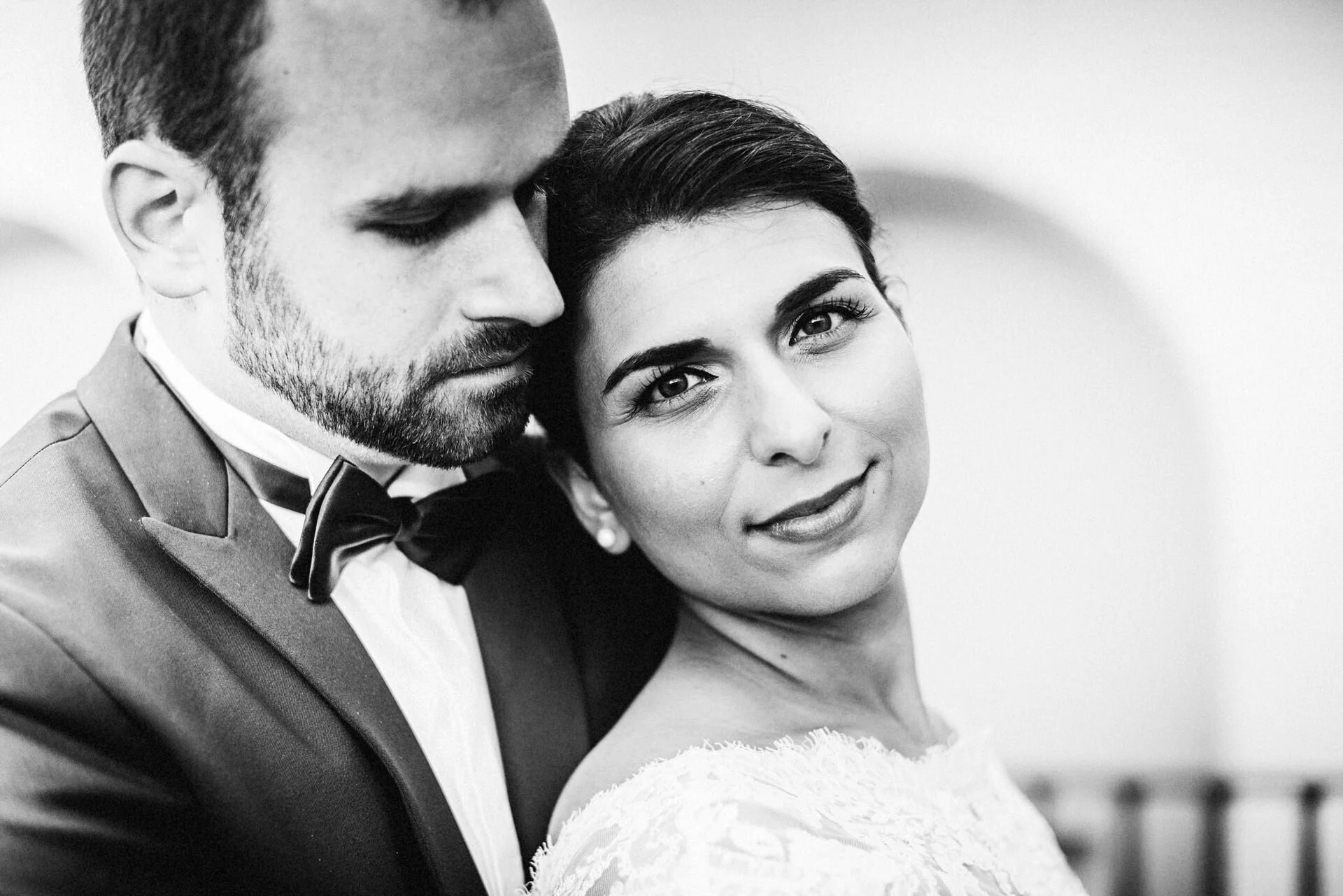Ein schwarz-weiß Portrait eines Paares an ihrem Hochzeitstag, das eng umeinander steht, der Mann trägt einen schwarzen Anzug und Fliege, die Frau ein weißes Spitzenkleid, beide schauen ins Kamera, die Frau lächelt leicht.