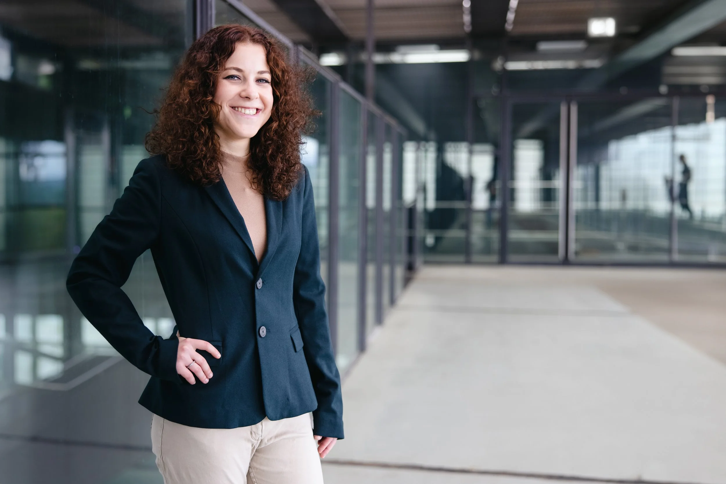 Eine lächelnde Frau in Business-Kleidung steht vor einem modernen Gebäude mit Glasfassade.