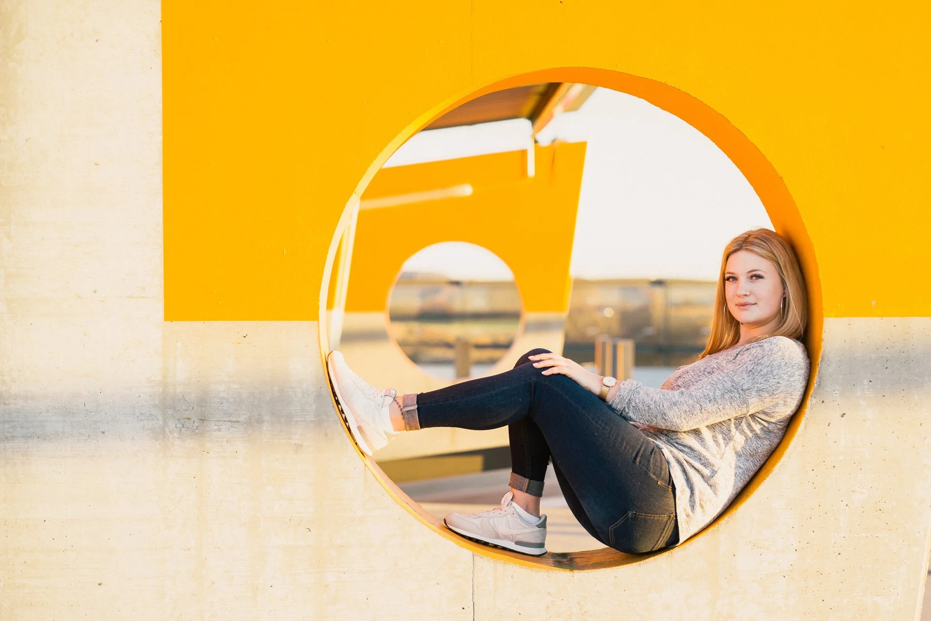 Junge Frau sitzt in einem großen gelben künstlerischen Rahmen im Freien, bei Sonnenuntergang, die Beine im Kreis ausgestreckt, lächelnd.