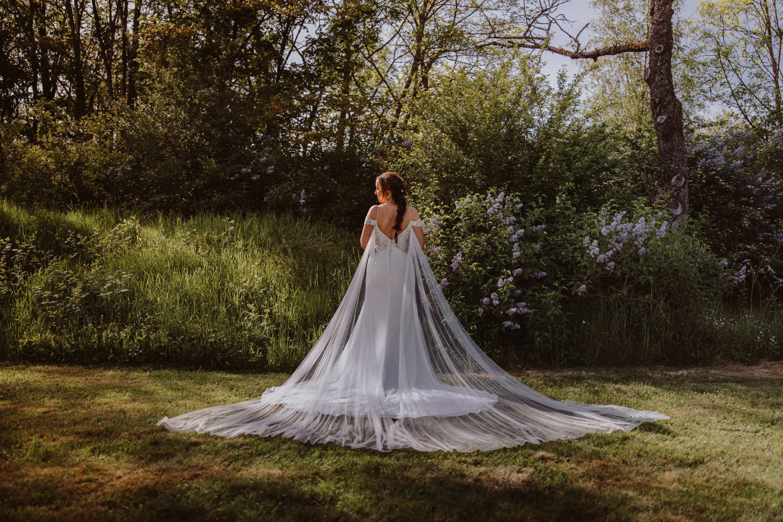 Braut in einem weißen Hochzeitskleid mit Schleppe steht im Garten, umgeben von Bäumen und blühenden Sträuchern, Sonnenlicht fällt durch die Blätter.