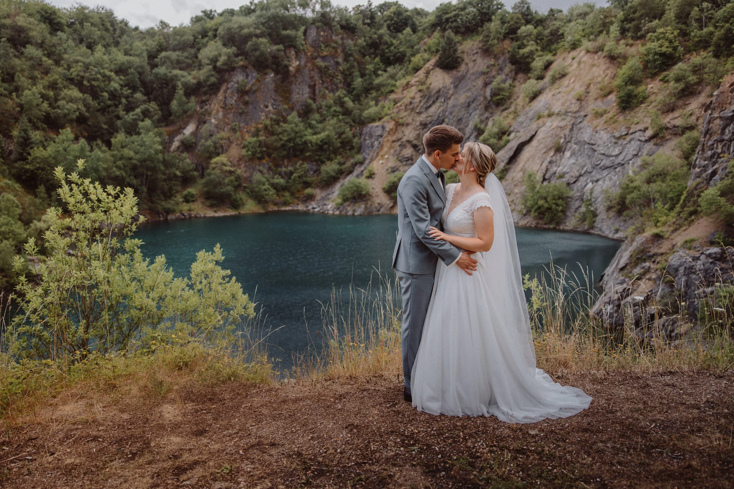 Ein Brautpaar in Hochzeitskleidung steht sich an einer natürlichen Wasserstelle umgeben von Felsen und Bäumen, wobei sie sich liebevoll zuhören.