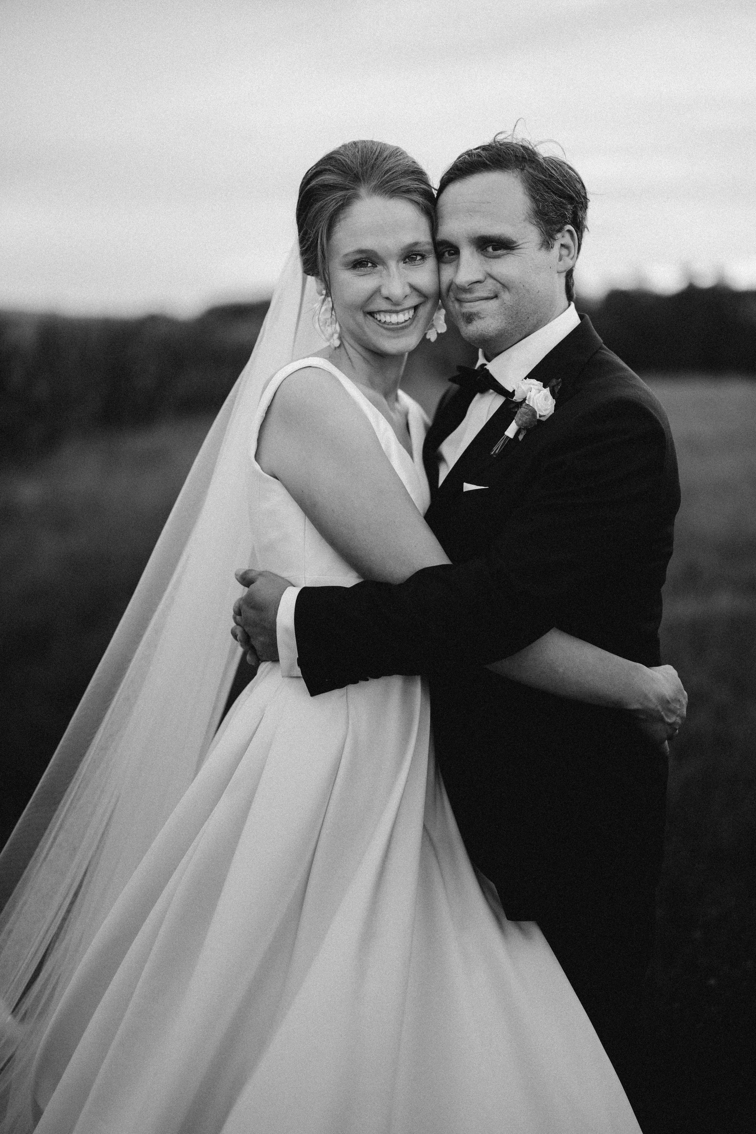 Ein Hochzeitsbild eines Paars in formeller Kleidung, ein Mann in einem schwarzen Anzug und eine Frau in einem weißen Hochzeitskleid mit Schleier, umarmen sich vor einer natürlichen Landschaft.