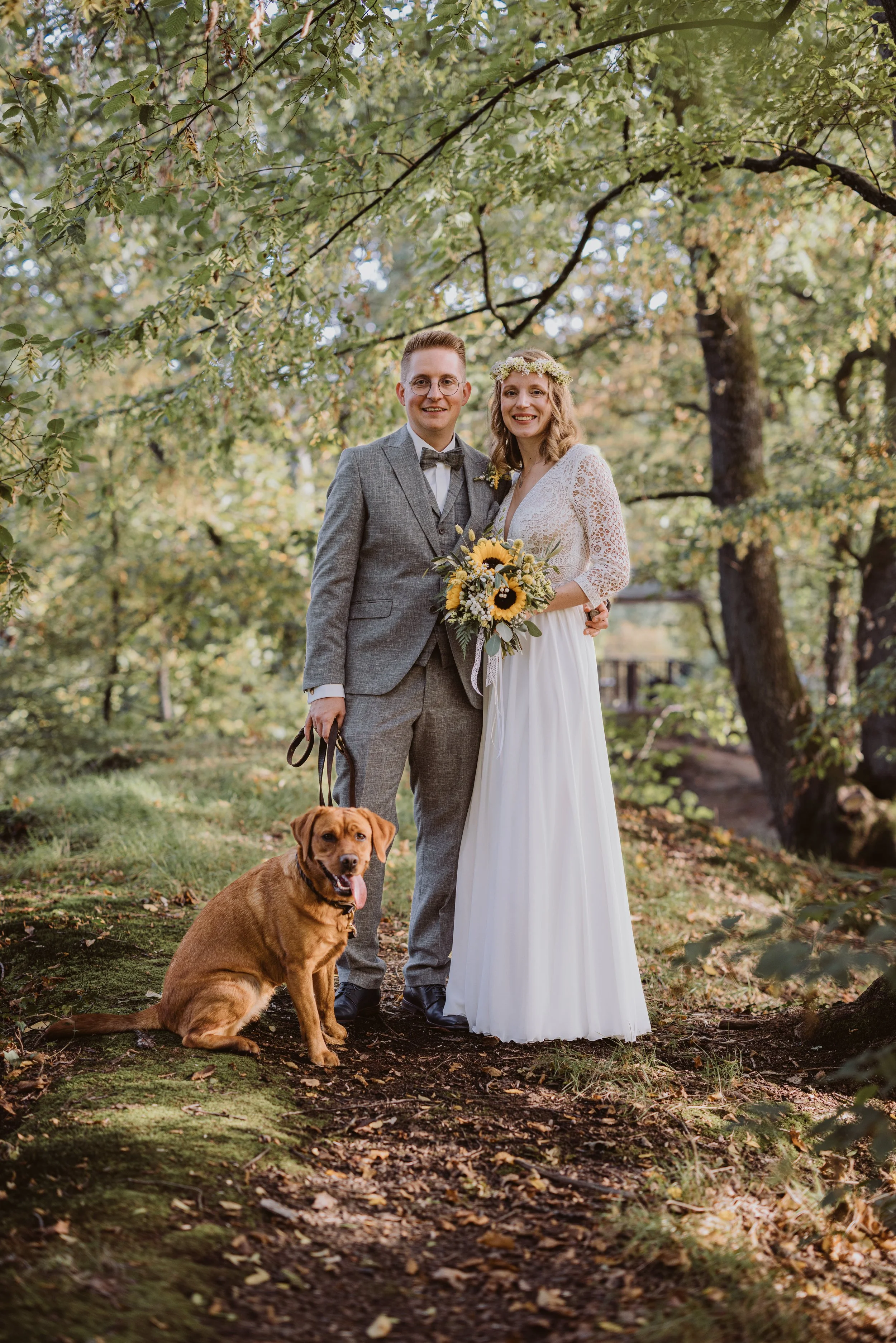 Ein brauner Hund sitzt neben einem lachenden Paar in formeller Kleidung, bei einer Hochzeitsaufnahme im Wald, mit viel Grün und Sonnenlicht im Hintergrund.
