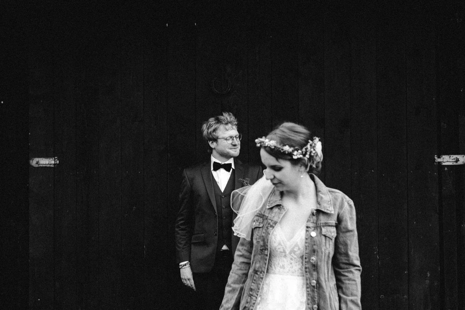 Schwarz-Weiß-Foto eines Paares bei einer Hochzeit, Frau trägt ein Kleid mit Fischgrätenärmel und Haar mit Blumenkranz, Mann trägt Anzug mit Fliege, hinter ihnen eine dunkle Holzwand.