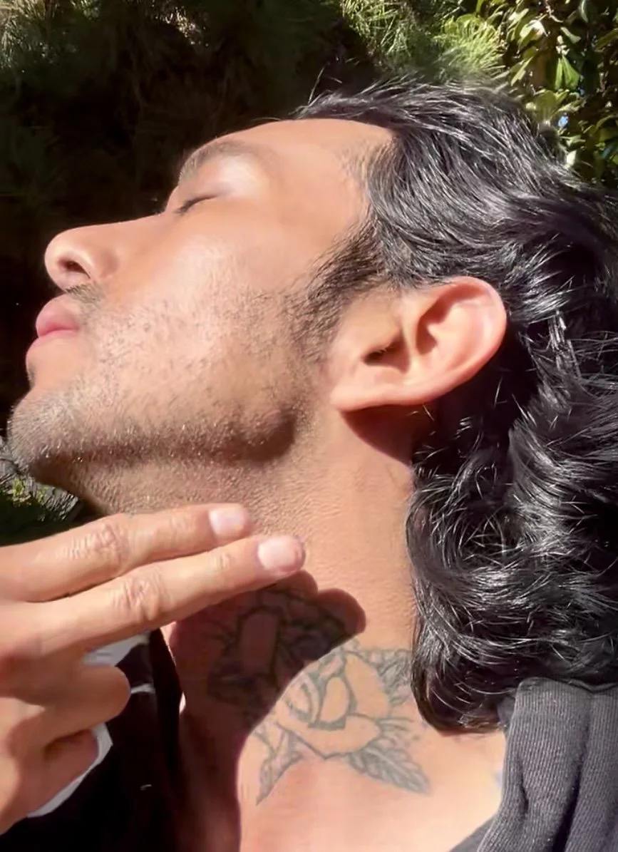 A man with long dark hair and a beard, touching his neck, with a tattoo of a rose on his shoulder, outdoors with trees in the background.