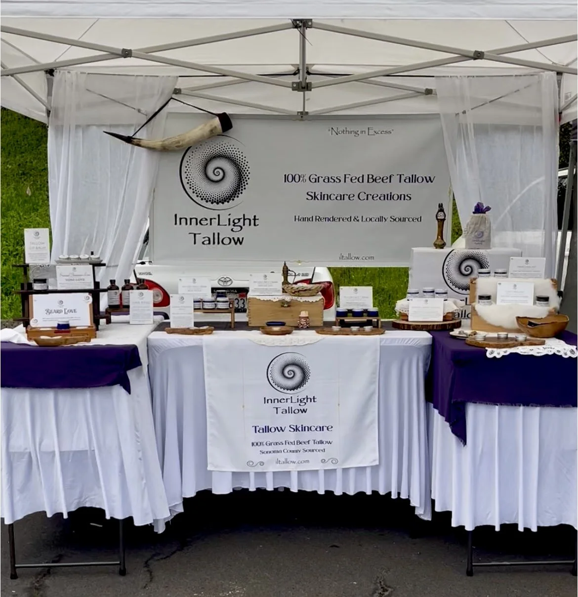 An outdoor market booth displaying skincare products made from grass-fed beef tallow. The booth is decorated with white and purple tablecloths, framed by white curtains, and features signs with the brand name Inner Light Tallow, slogans, and product information. Various jars and containers of skincare creams are arranged on the tables.