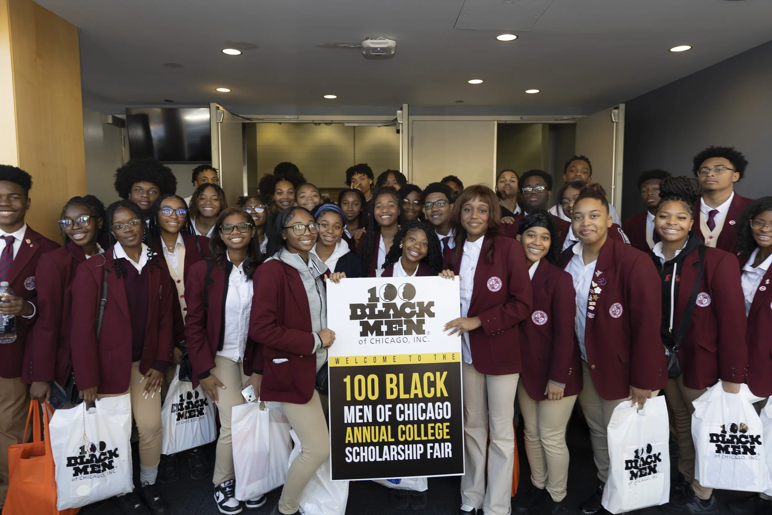 Group of students wearing maroon blazers at a college scholarship fair, holding a sign that reads 'Welcome to the 100 Black Men of Chicago, Inc. Annual College Scholarship Fair.'