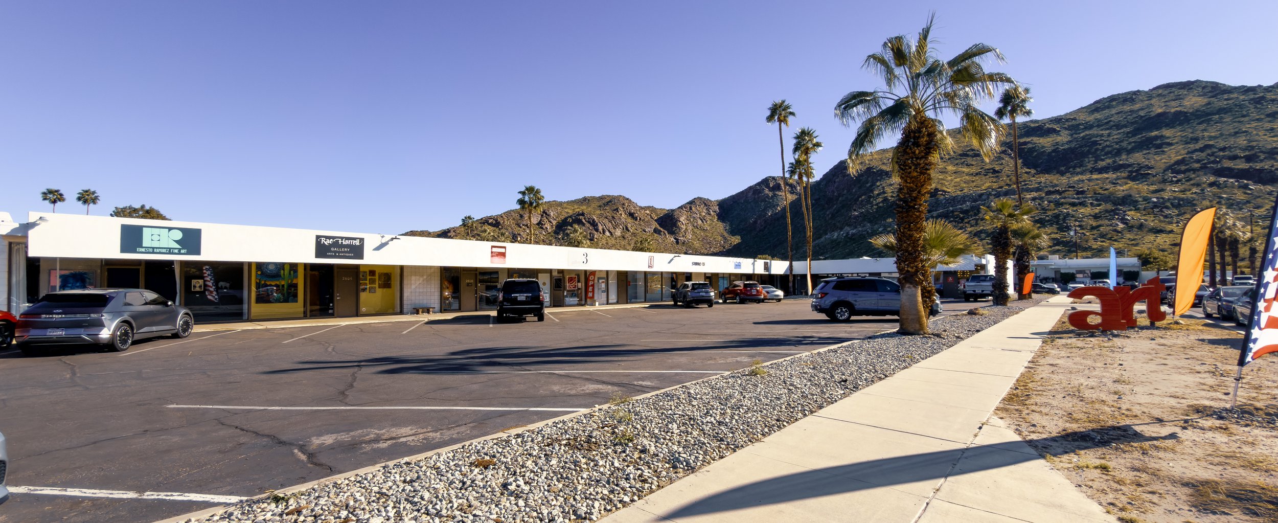 Commercial strip mall with art gallery, parked cars, palm trees, and mountains in the background during daytime.