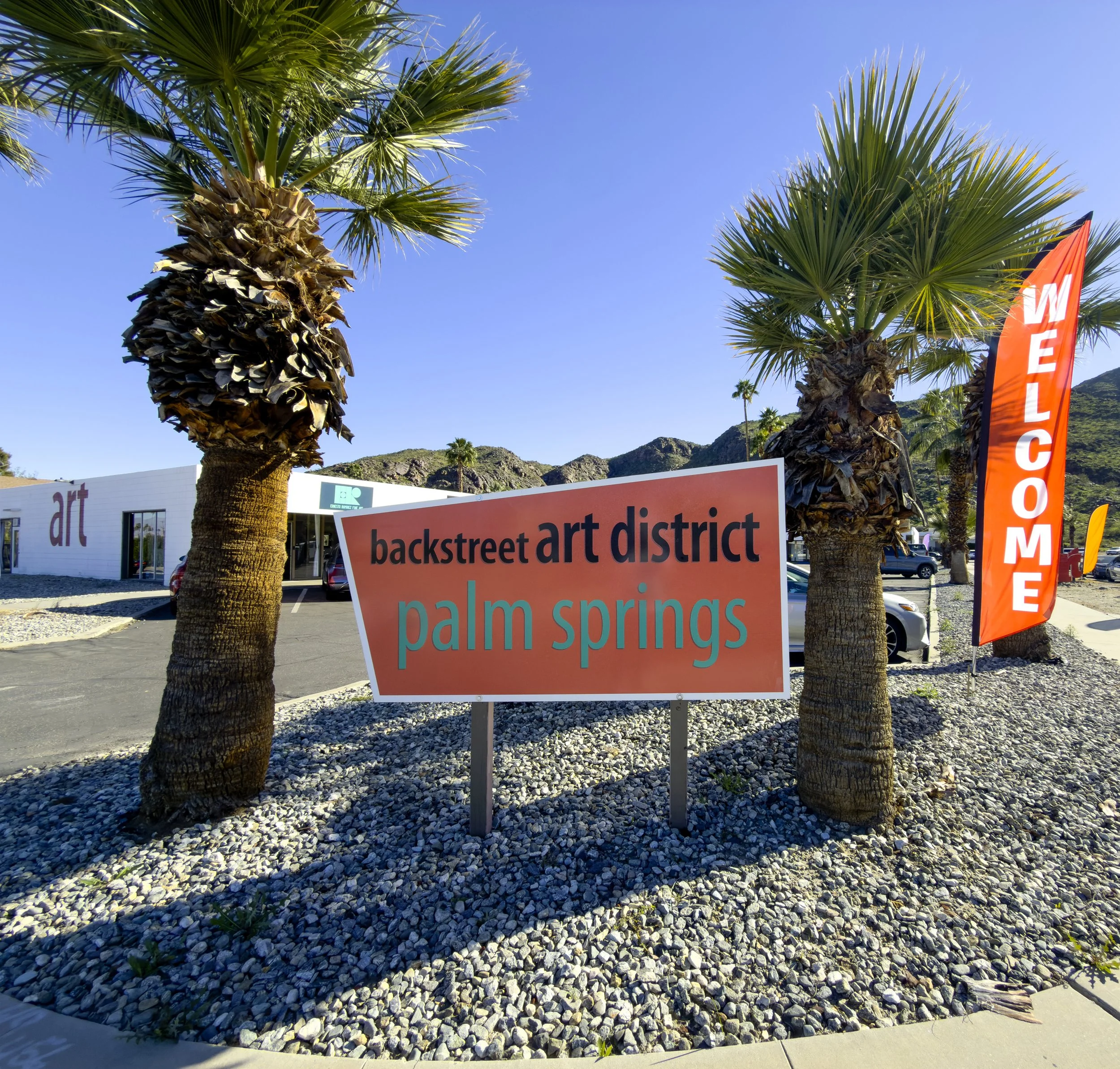 Sign reading 'backstreet art district palm springs' with palm trees and a building in the background, and a red 'welcome' banner to the right.