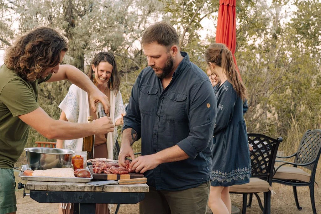 People in a kitchen preparing food, with a man in a tank top working at a kitchen island, surrounded by women observing. The island is topped with jars and a tray of small dishes. The kitchen features wooden cabinetry and a large sink by the window.