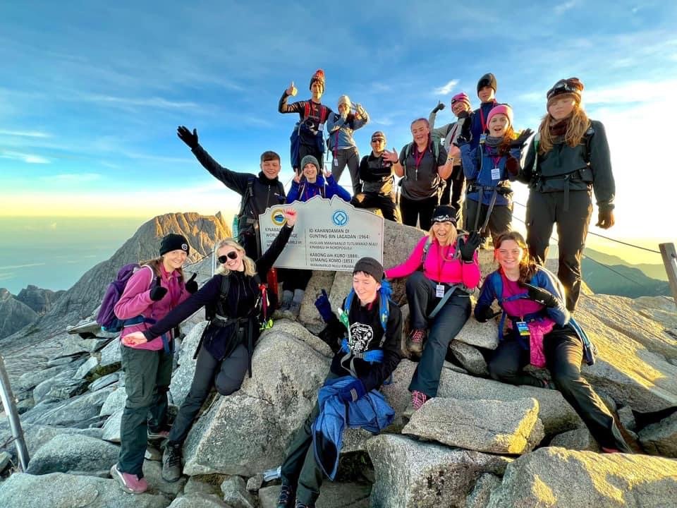 A school group celebrating at the summit of Mount Kinabalu in Borneo at sunrise, gathered around the official summit plaque at 4,095 metres.