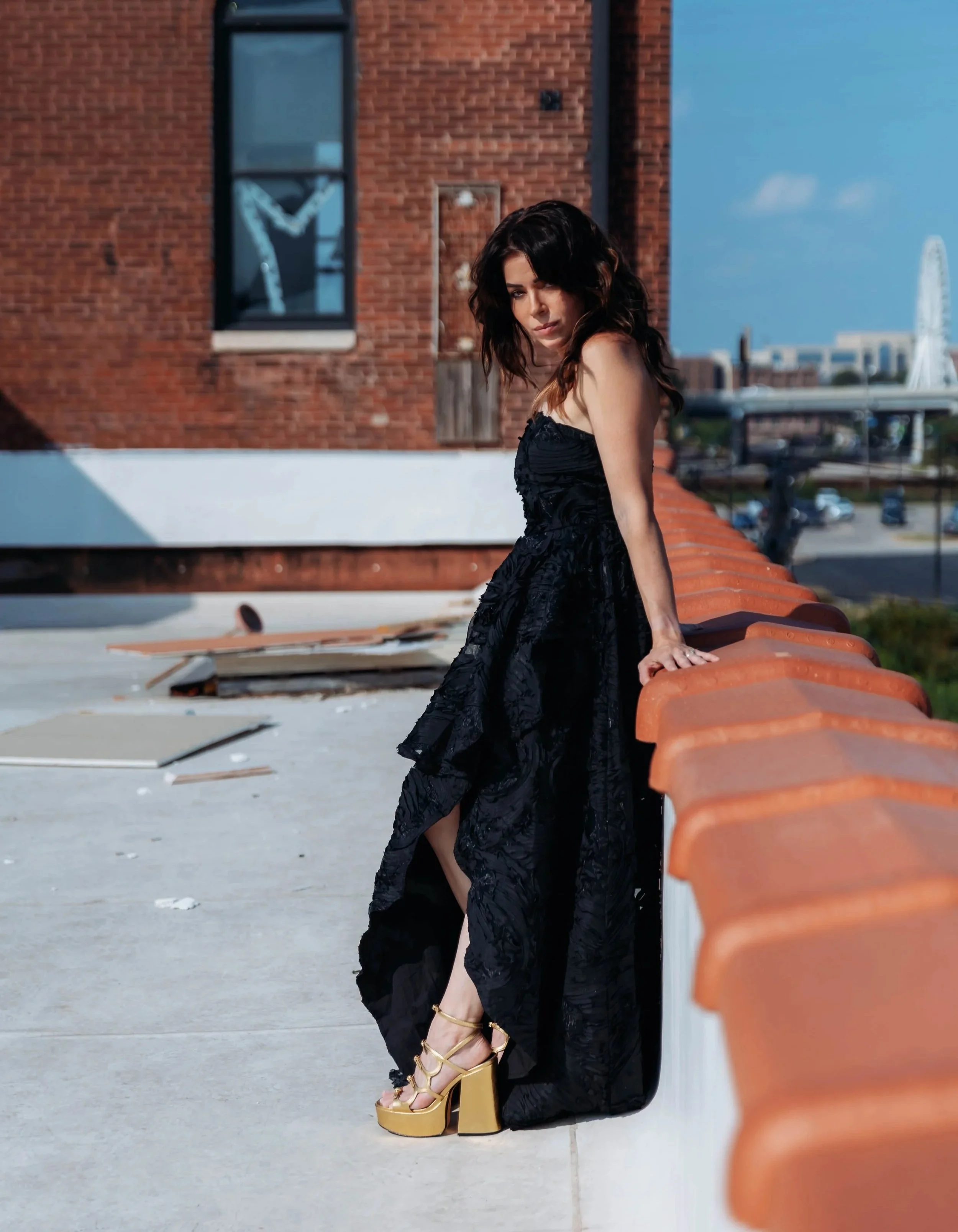 A woman wearing a long black dress and gold platform high heels standing on a rooftop with a brick building, a city skyline in the background.