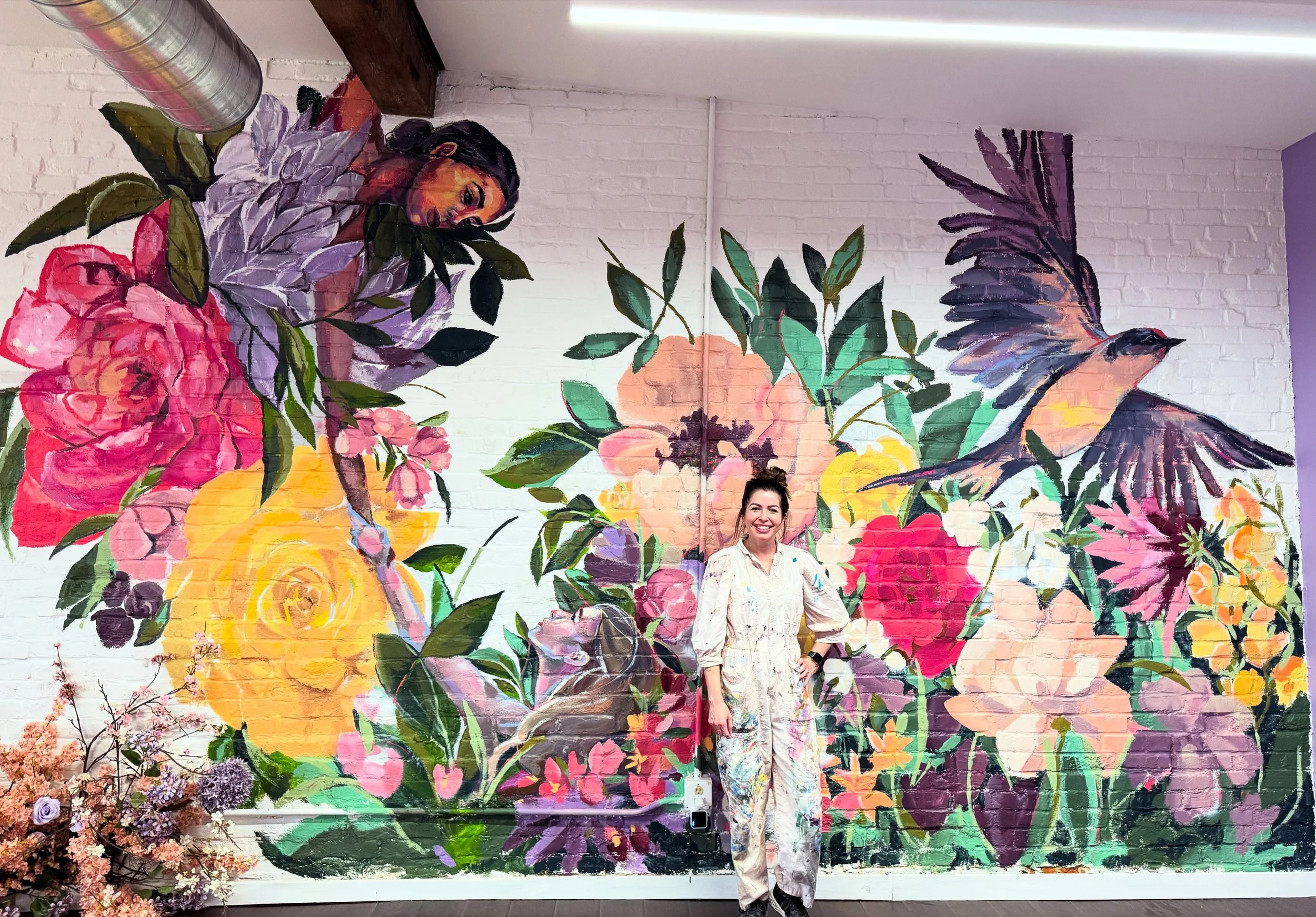 Elysia King standing in front of a mural of a woman leaning down towards flowers, foliage, and a hummingbird