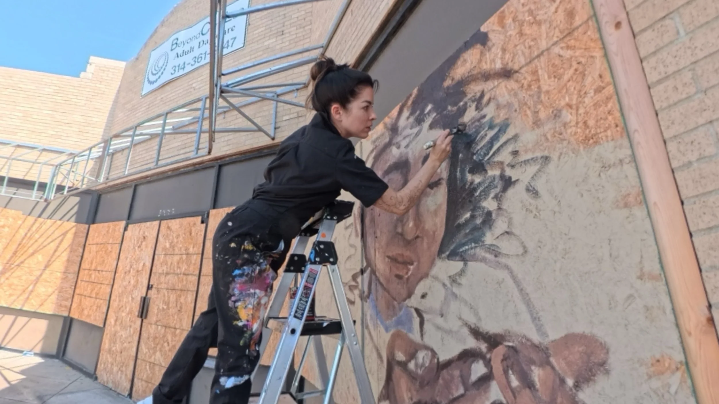 Elysia King on a ladder painting a mural of a woman outdoors