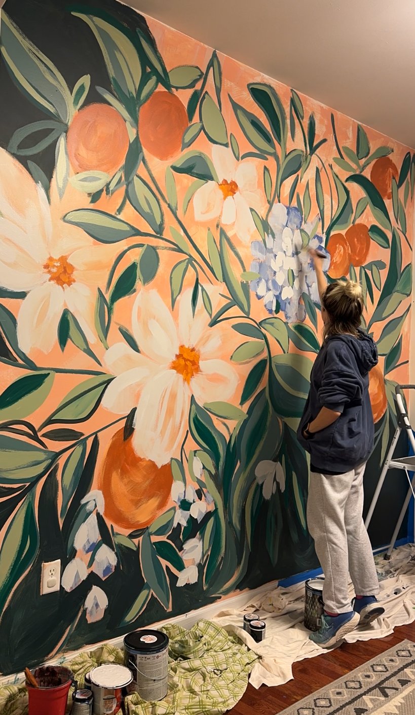 Elysia painting a mural of oranges, leaves, and white flowers against a salmon pink background